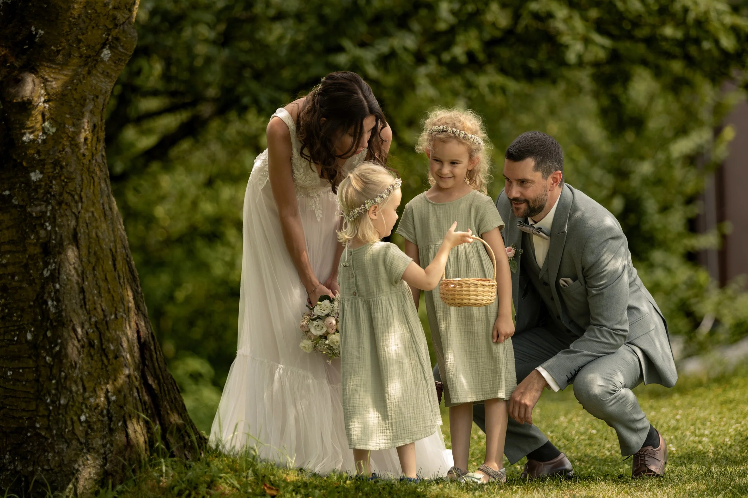 Eine Frau im weißen Kleid, ein Mann im grauen Anzug, zwei kleine Mädchen mit Blumenkränzen im Haar, stehen im Grünen unter einem Baum bei einer Hochzeit im Freien.