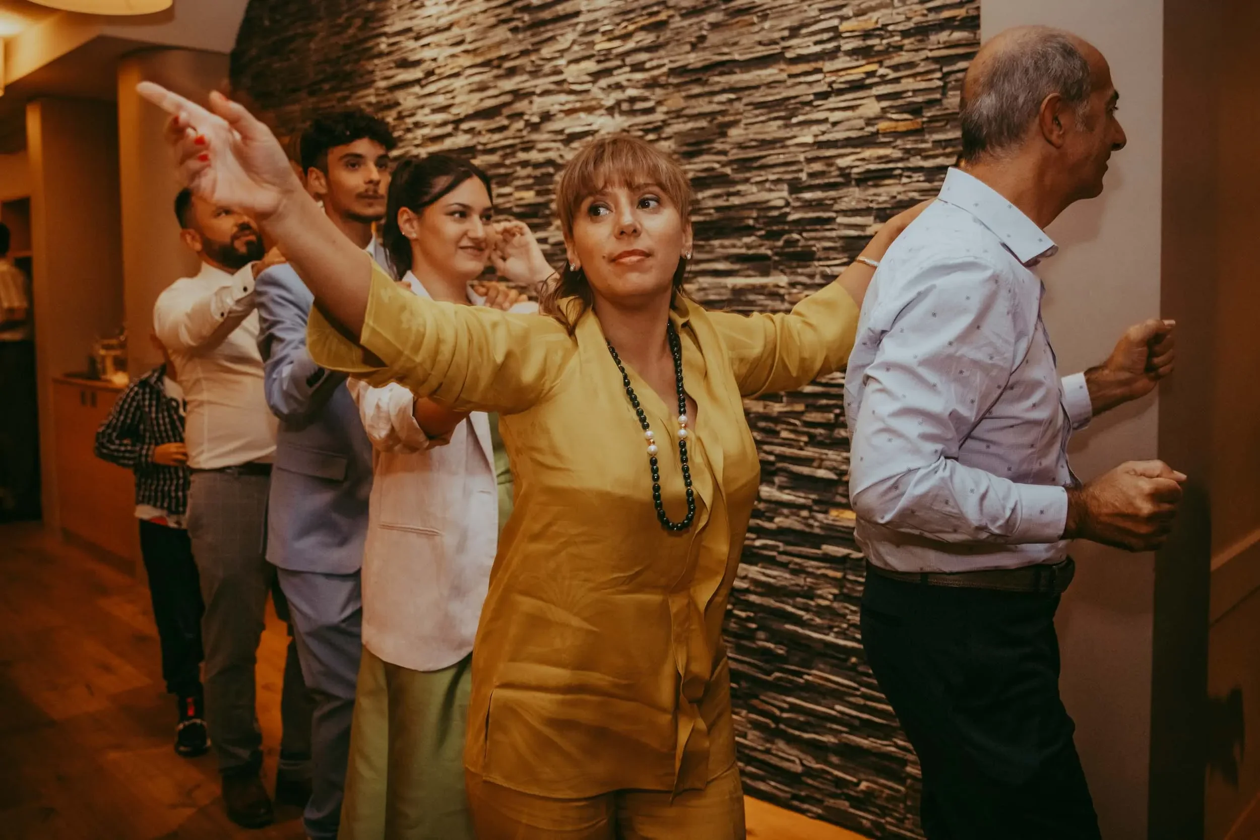 Fünf Menschen stehen in einer Reihe in einem Raum mit einer Steinwand im Hintergrund, alle sind in einer Linie ausgerichtet und haben die Arme auf den Schultern der Person vor ihnen gelegt.