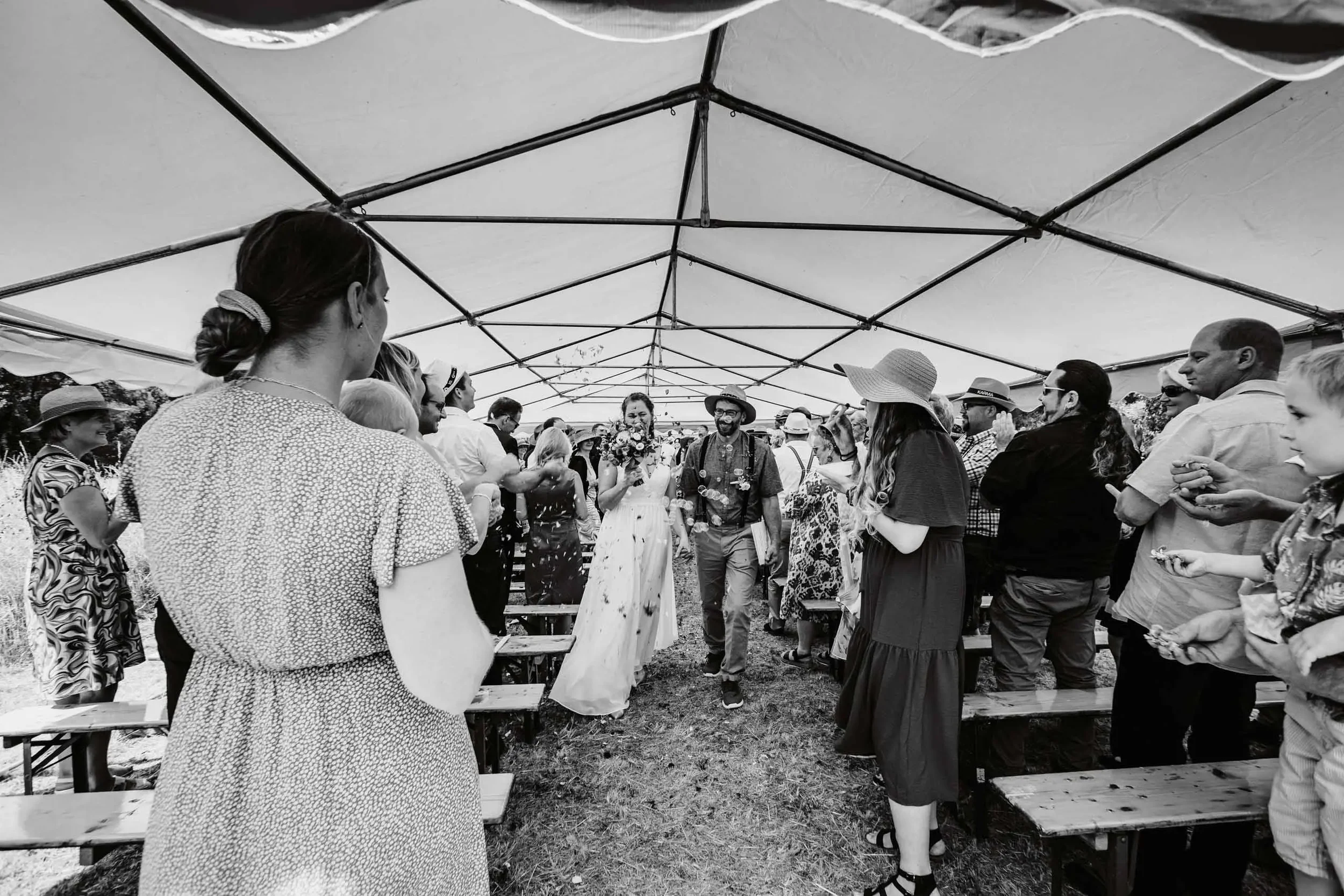 Menschen feiern unter einem großen Zelt, einige sind lachend und trinken, others tragen Hüte und Sommerkleidung.
