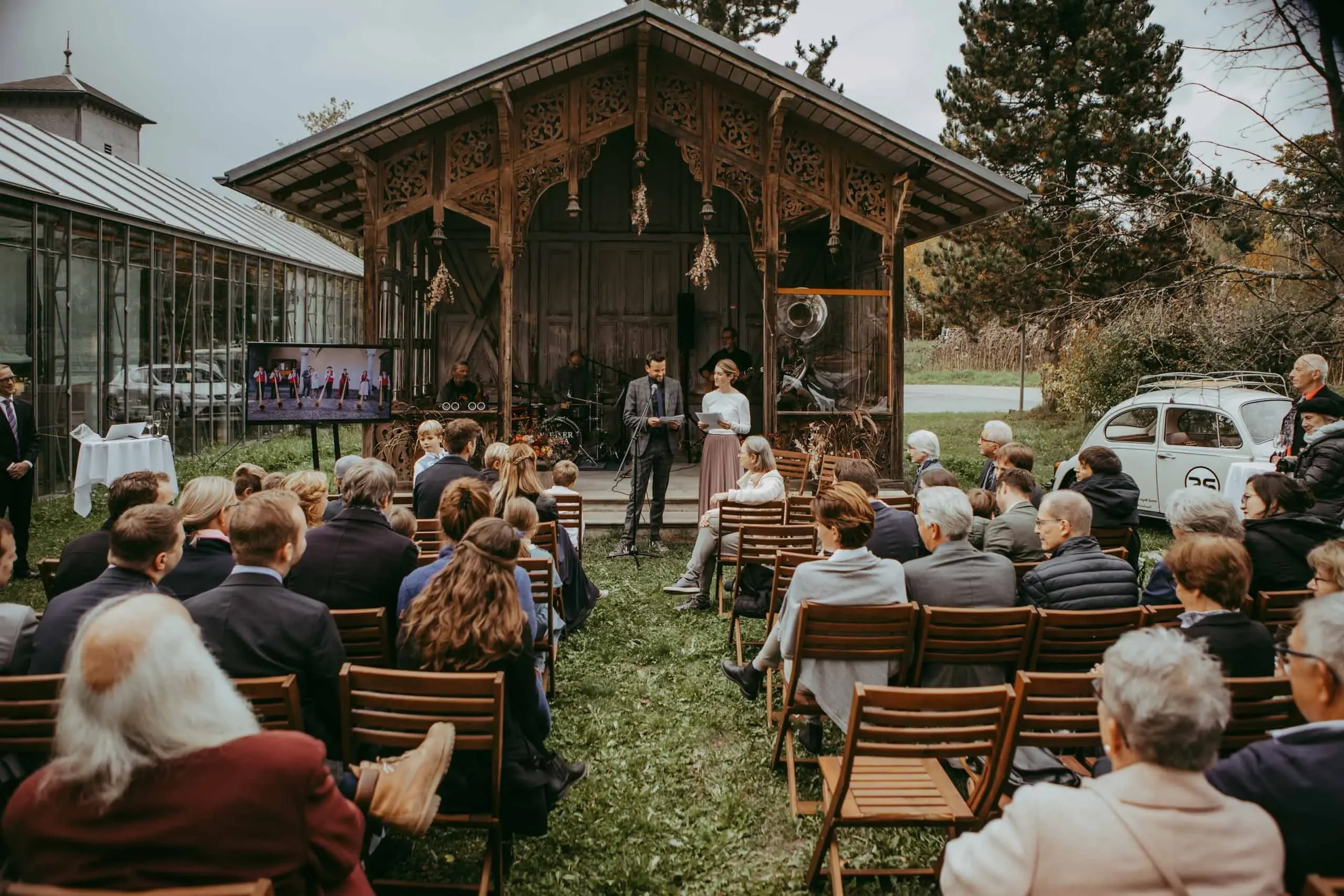 Menschen bei einer Outdoor-Veranstaltung, die auf einer Bühne mit Holzverzierung sprechen oder aufführen, während sie von einem Publikum aus sitzenden Personen beobachtet werden.