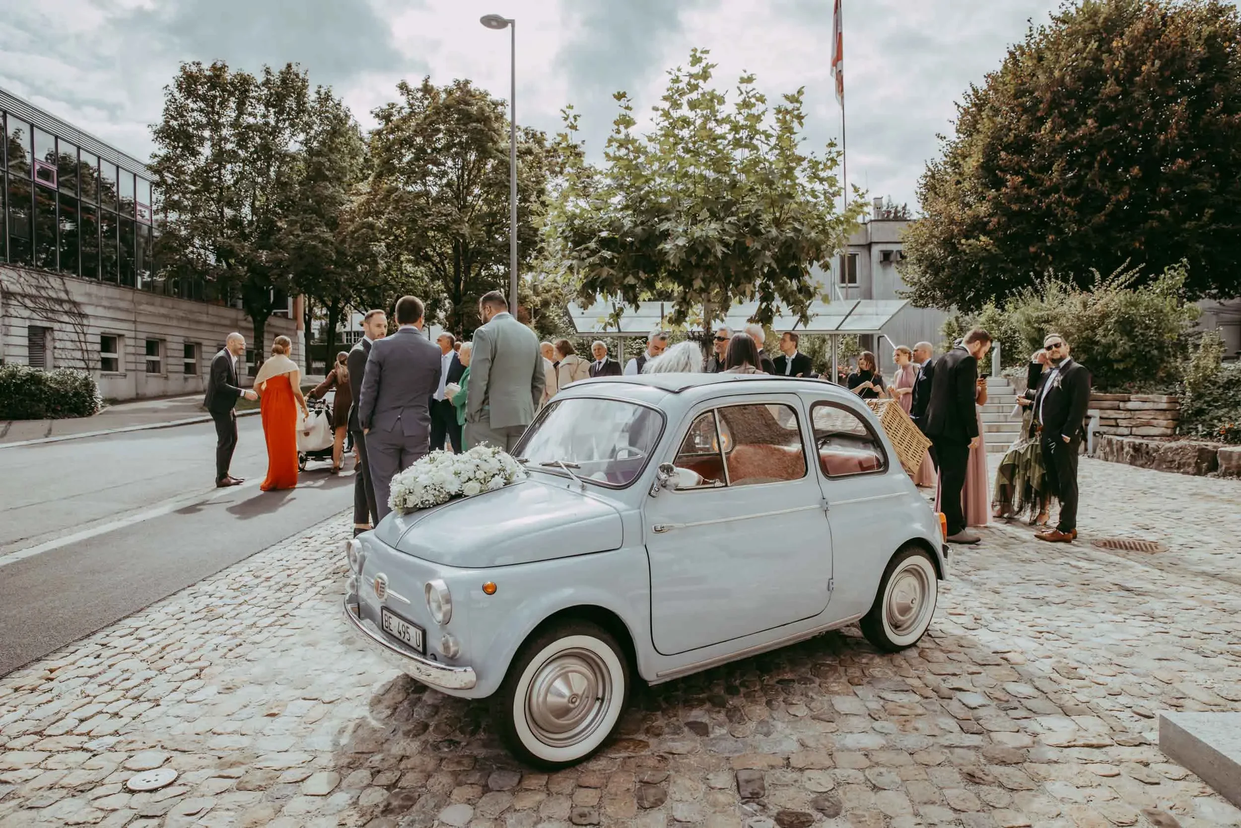 Eine Gruppe von Menschen in formeller Kleidung, wahrscheinlich bei einer Hochzeit, steht vor einem modernen Gebäude im Freien, neben einem alten weißen Fiat 500 mit Blumen auf der Motorhaube.