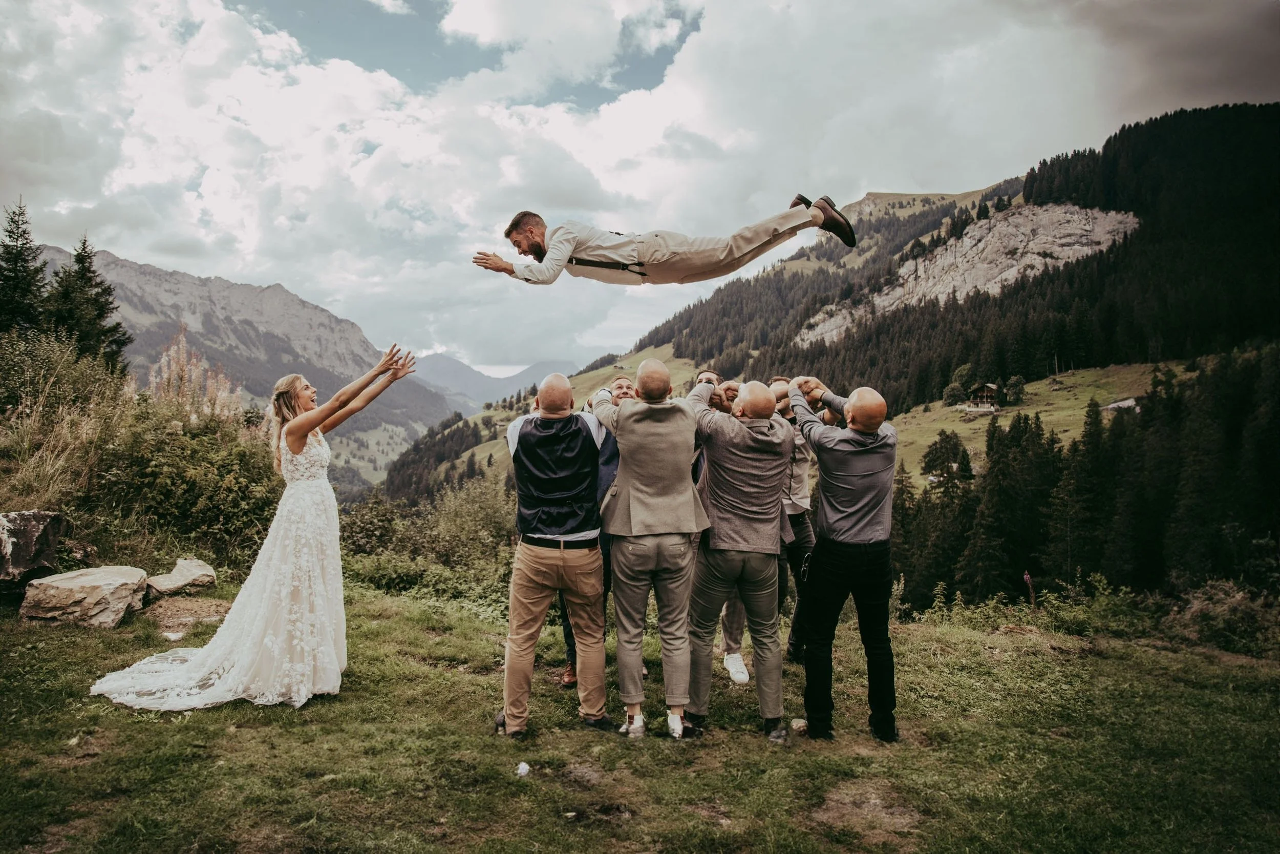 Ein Mann in Anzug liegt auf dem Rücken in der Luft, während eine Gruppe von Männern ihn hochhält. Eine Frau in Hochzeitskleid steht neben der Gruppe. Die Szene findet im Freien in einer bergigen Landschaft statt.
