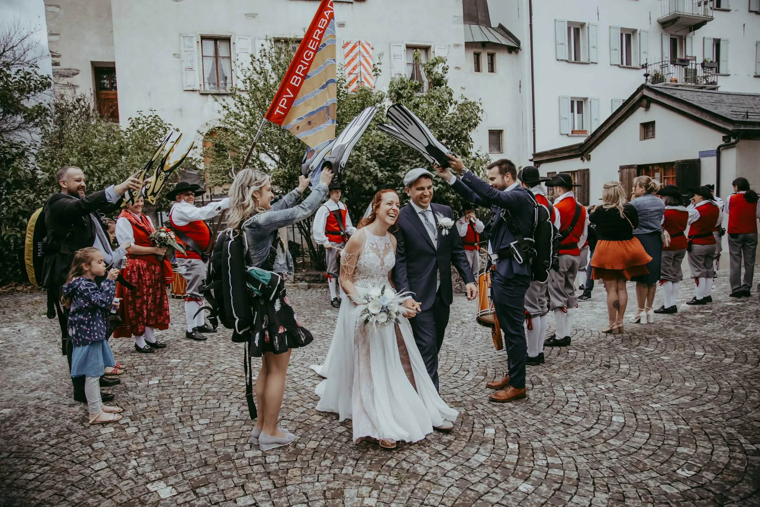 Hochzeitspaar feiern, umgeben von Menschen in traditioneller Kleidung, auf einer gepflasterten Straße in einer Stadt mit weißen Gebäuden und mehreren Fenstern und Balkonen.