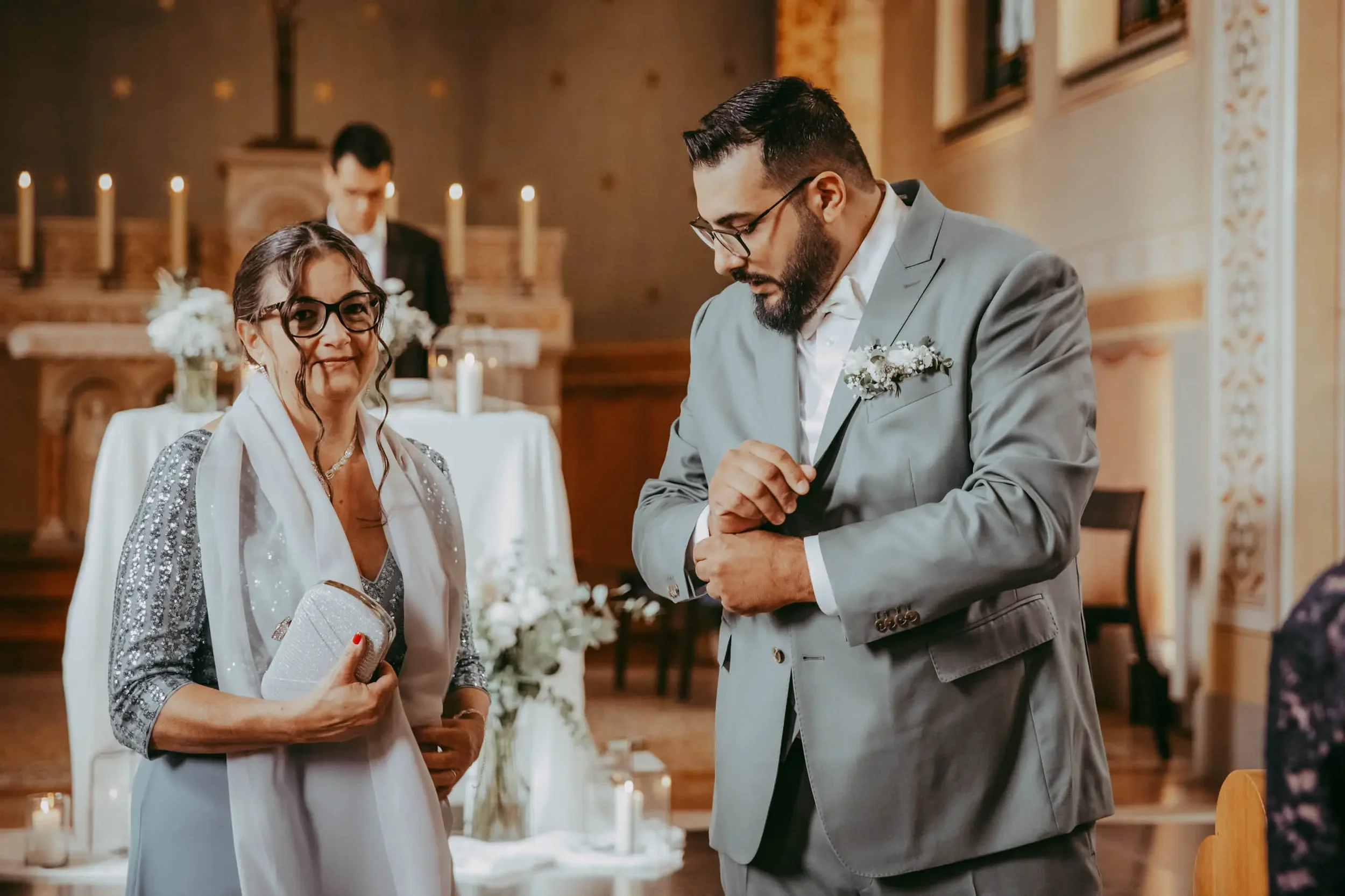 Ein Mann in einem grauen Anzug ringt während einer Hochzeitszeremonie mit seiner Uhr, während eine Frau in einem silbernen Kleid und großen Brillen schminkt und lächelt.