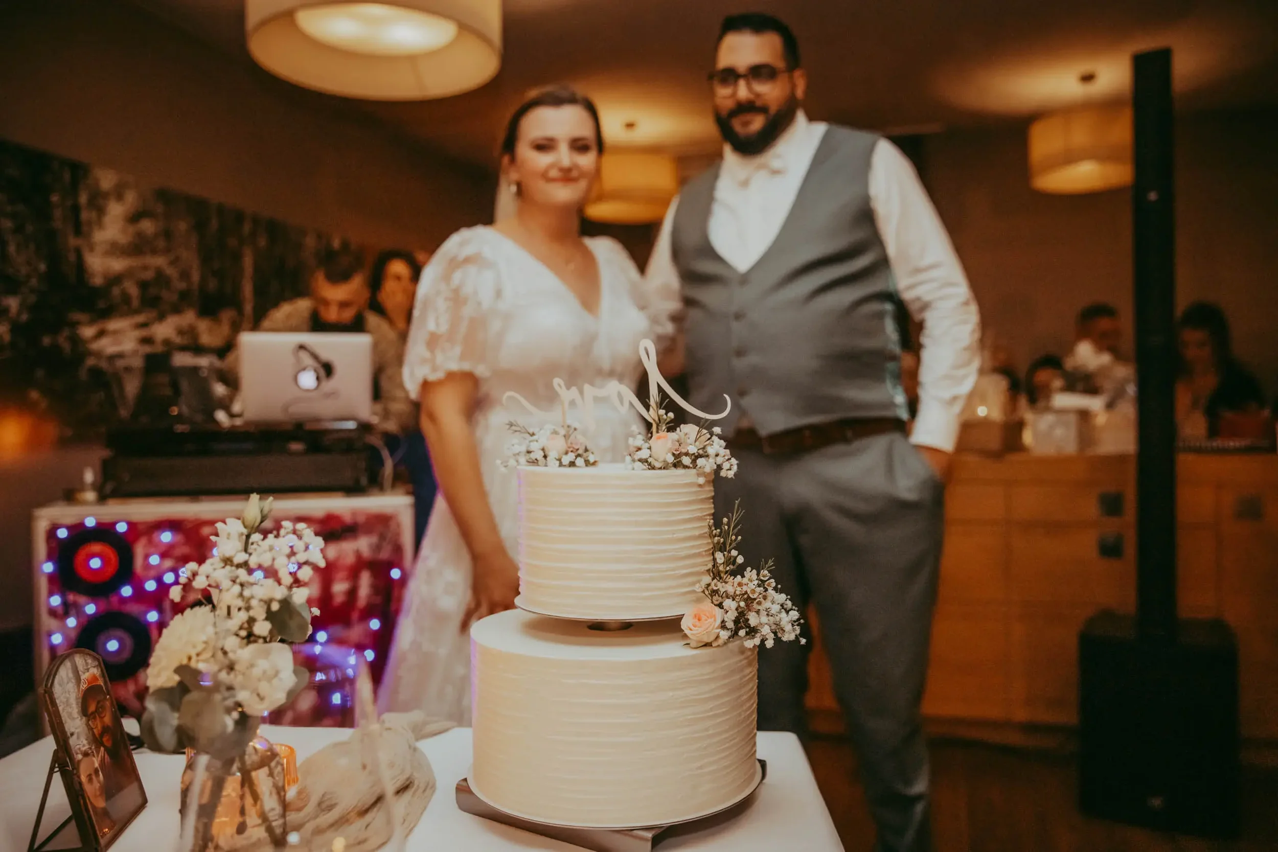 Ein Brautpaar bei ihrer Hochzeit, stehend hinter einer großen, weißen, gestuften Hochzeitstorte mit Blumen. Im Hintergrund sind Gäste, eine DJ-Anlage mit Lichtern, und ein DJ, der an einem Laptop arbeitet. Die Atmosphäre ist warm und festlich.