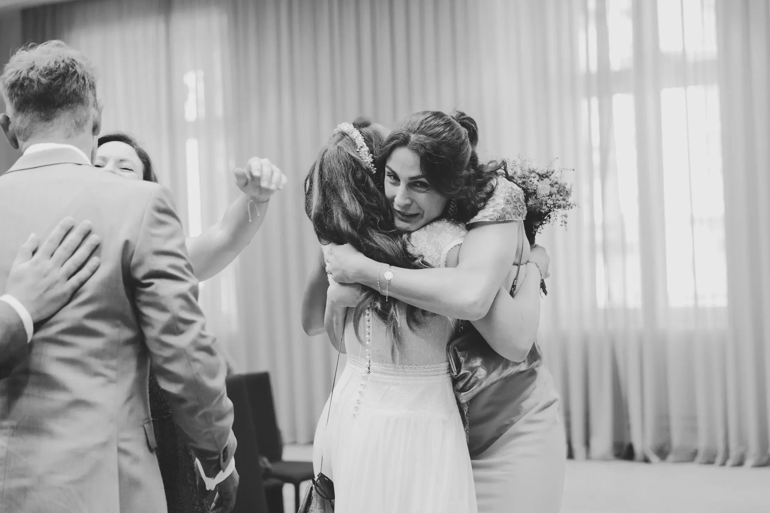 Zwei Frauen umarmen sich bei einer Hochzeit, während sie trösten und freudig sind, in einem Raum mit großen Fenstern und Vorhängen.