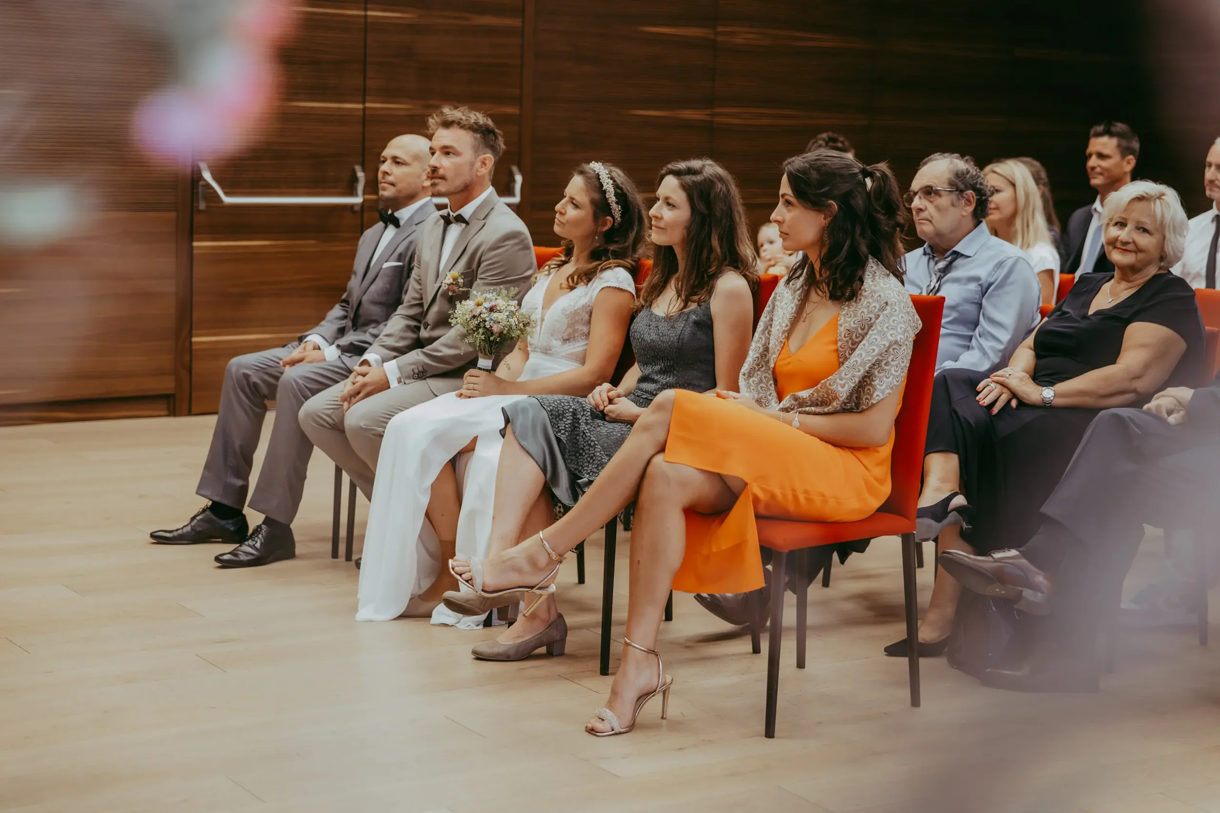 Eine Gruppe von Gästen bei einer Hochzeitsfeier sitzt in einem Saal, der auf eine formelle Veranstaltung wie eine Hochzeit hindeutet.