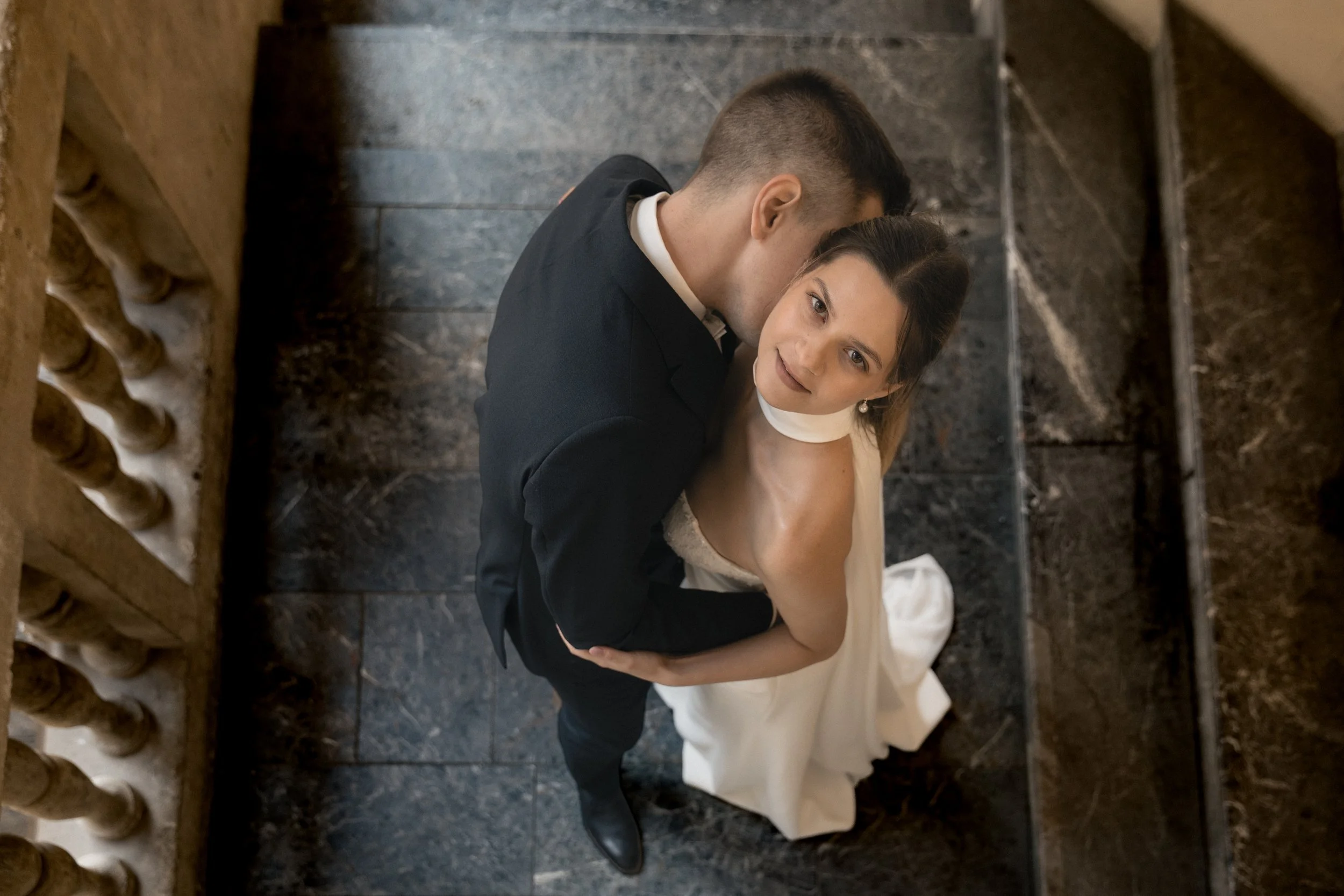 Ein Mann und eine Frau sitzen eng umarmt auf einer Treppe aus dunklem Stein. Der Mann trägt einen schwarzen Anzug und die Frau ein weißes Kleid. Sie schauen sich liebevoll an.