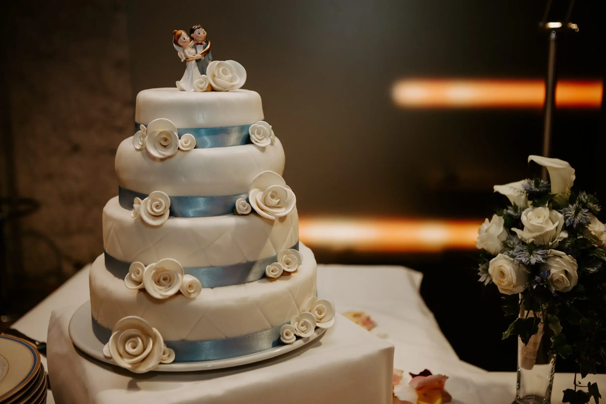 Mehrstöckige Hochzeitstorte mit weißen Rosen und Hasenfigur auf der Oberseite, daneben ein Blumenstrauß.