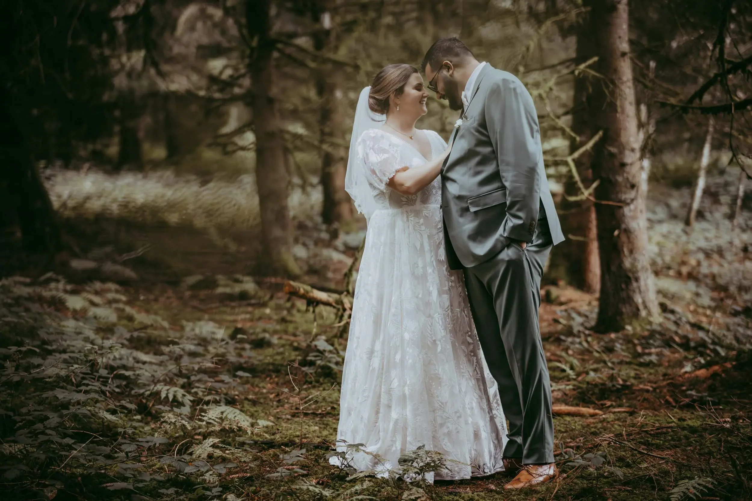 Ein Brautpaar steht im Wald, die Frau trägt ein weißes Hochzeitskleid, der Mann einen grauen Anzug, beide lächeln sich an und stehen sehr nah beieinander.