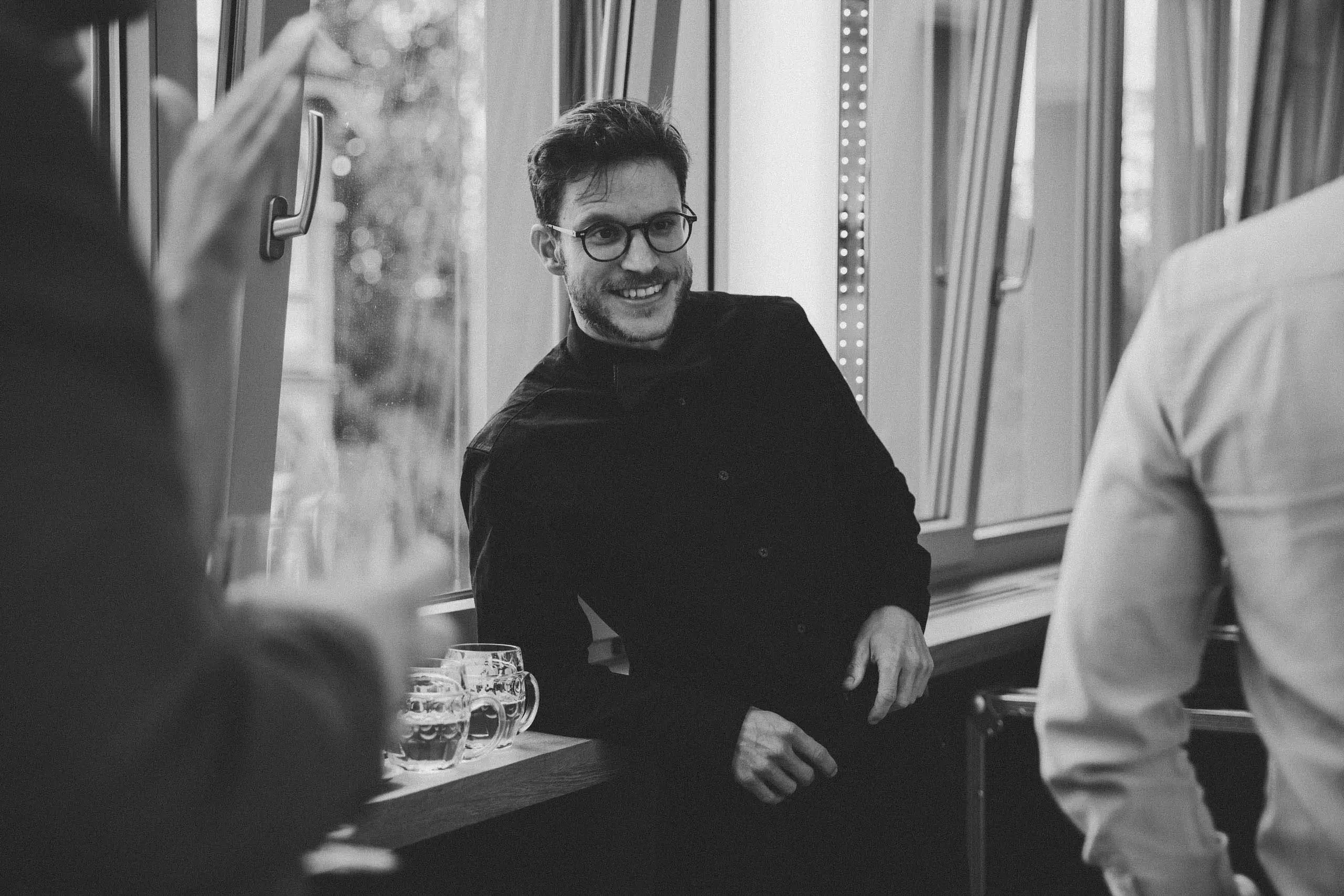 Ein Mann mit Brille und dunklem Hemd sitzt lächelnd in einem Raum mit großen Fenstern, während andere Menschen sich unterhalten, und einige Bier gläser stehen auf einem Tisch.