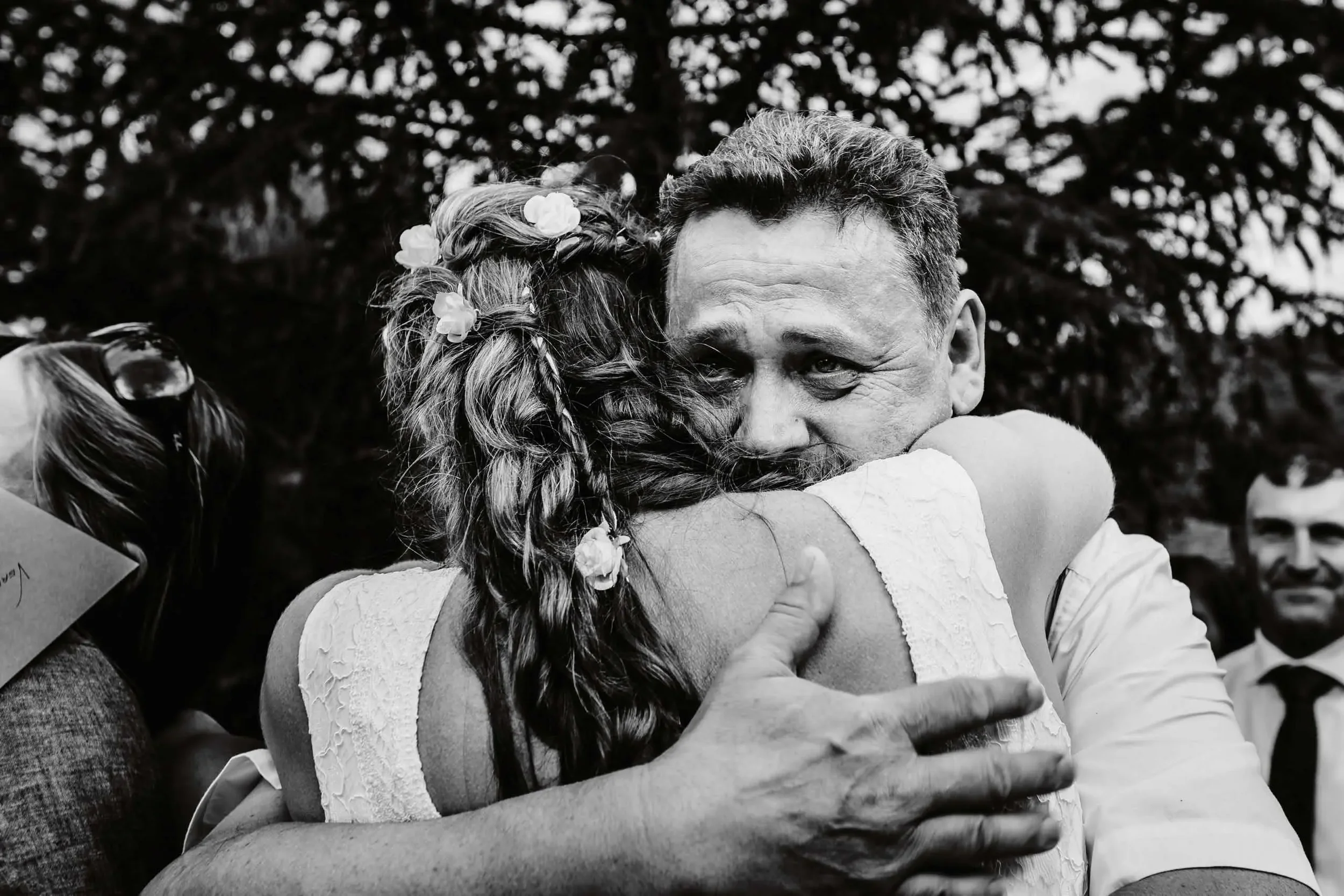 Zwei Menschen umarmen sich eng, Freude und Emotionen sichtbar, im Hintergrund unscharfer Park oder Garten mit Bäumen, schwarz-weiß Fotografie.