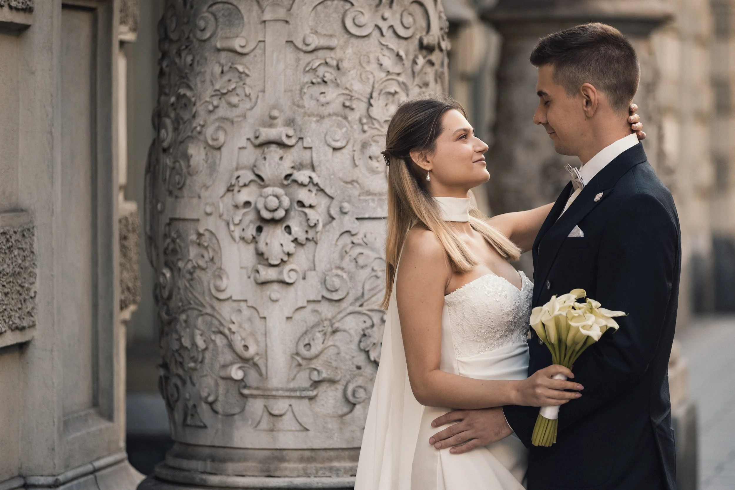 Ein Brautpaar bei einer Hochzeit steht sich gegenüber, eine Frau in einem weißen Hochzeitskleid und ein Mann in einem schwarzen Anzug mit Fliege. Die Frau hält einen Blumenstrauß aus weißen Calla-Lilien. Sie schauen sich liebevoll an, vor einer histo