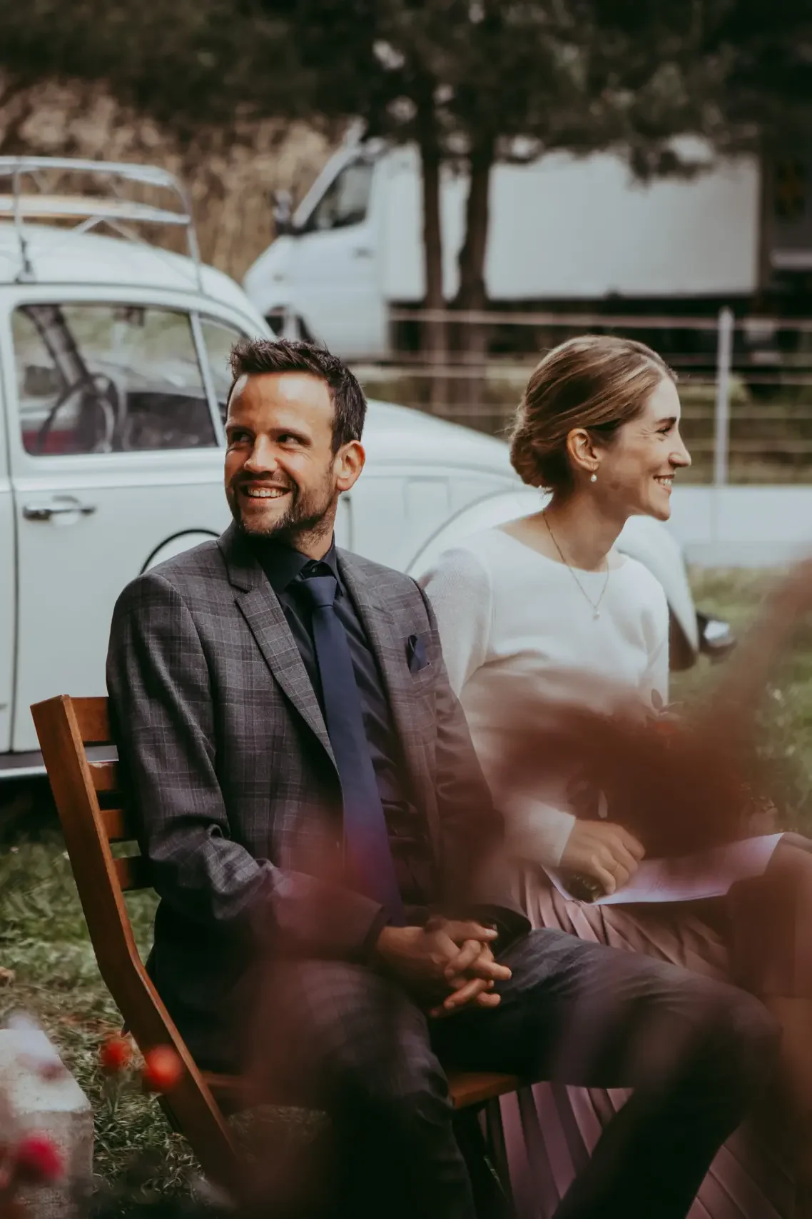 Ein Mann und eine Frau sitzen nebeneinander im Freien auf einem Klappstuhl. Der Mann trägt einen grauen Anzug mit Karo-Muster und eine dunkle Krawatte, die Frau trägt eine weiße Bluse. Im Hintergrund sind zwei weiße Fahrzeuge und Bäume sichtbar.