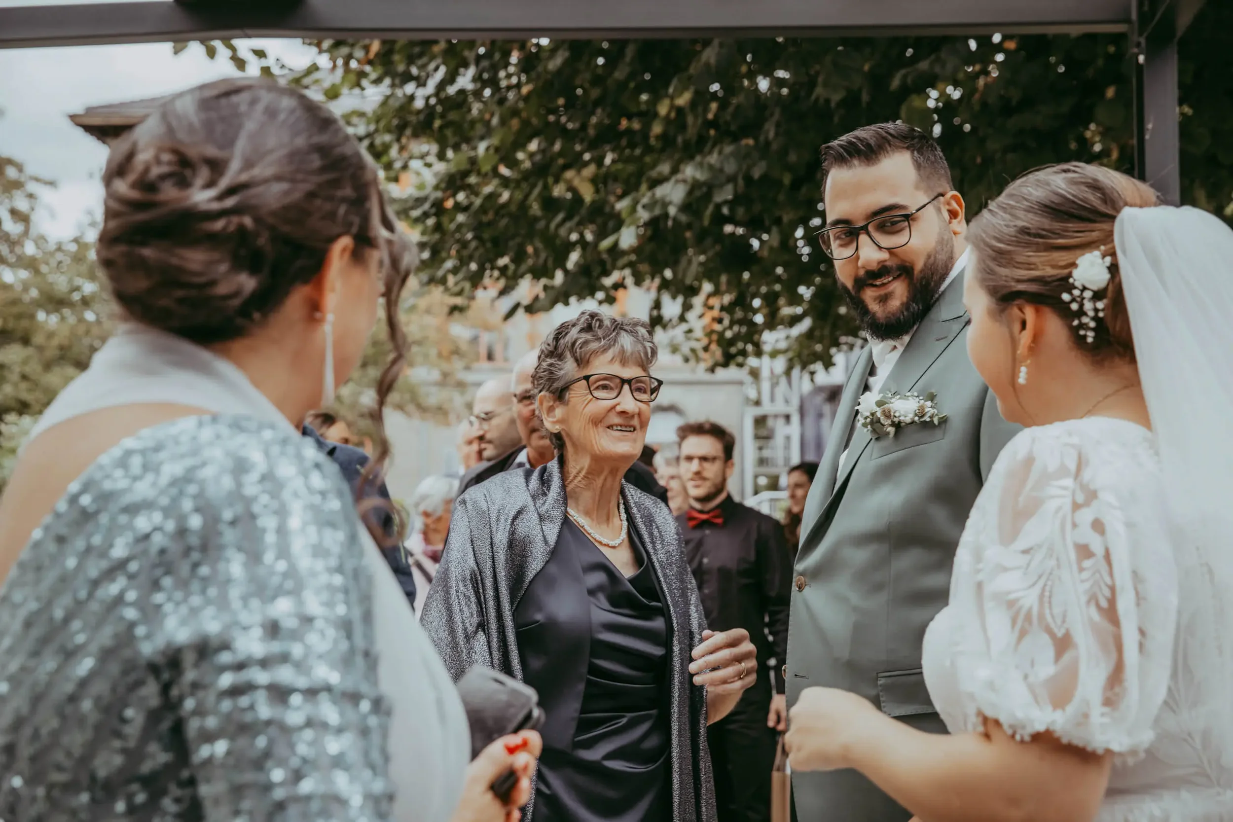 Hochzeitspaar und ältere Frau beim Händeschütteln unter einem Baum im Freien.