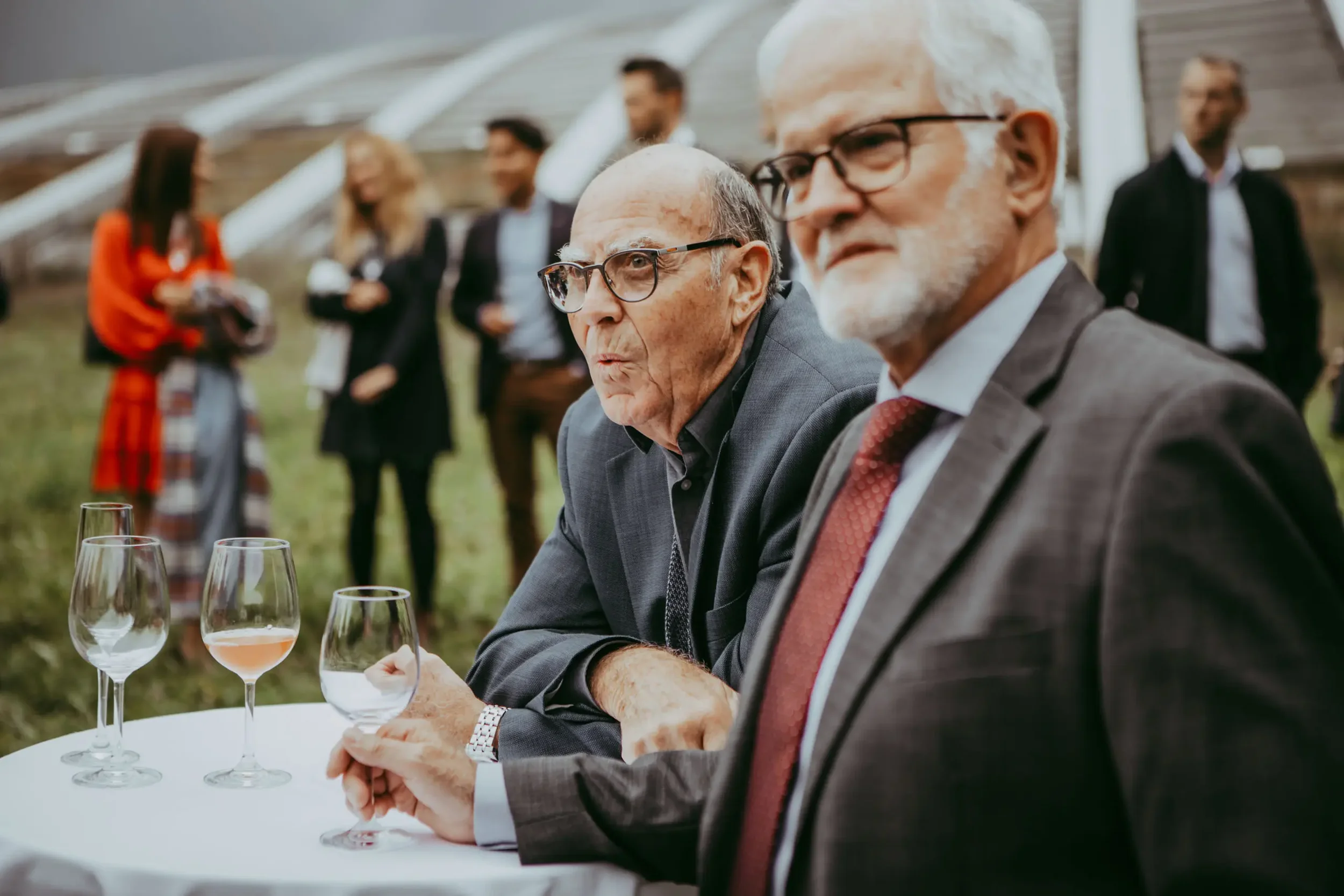 Älterer Mann mit Brille und Anzug sitzt bei einer Weinverkostung im Freien, im Hintergrund weitere Gäste beim Gespräch und Wein.