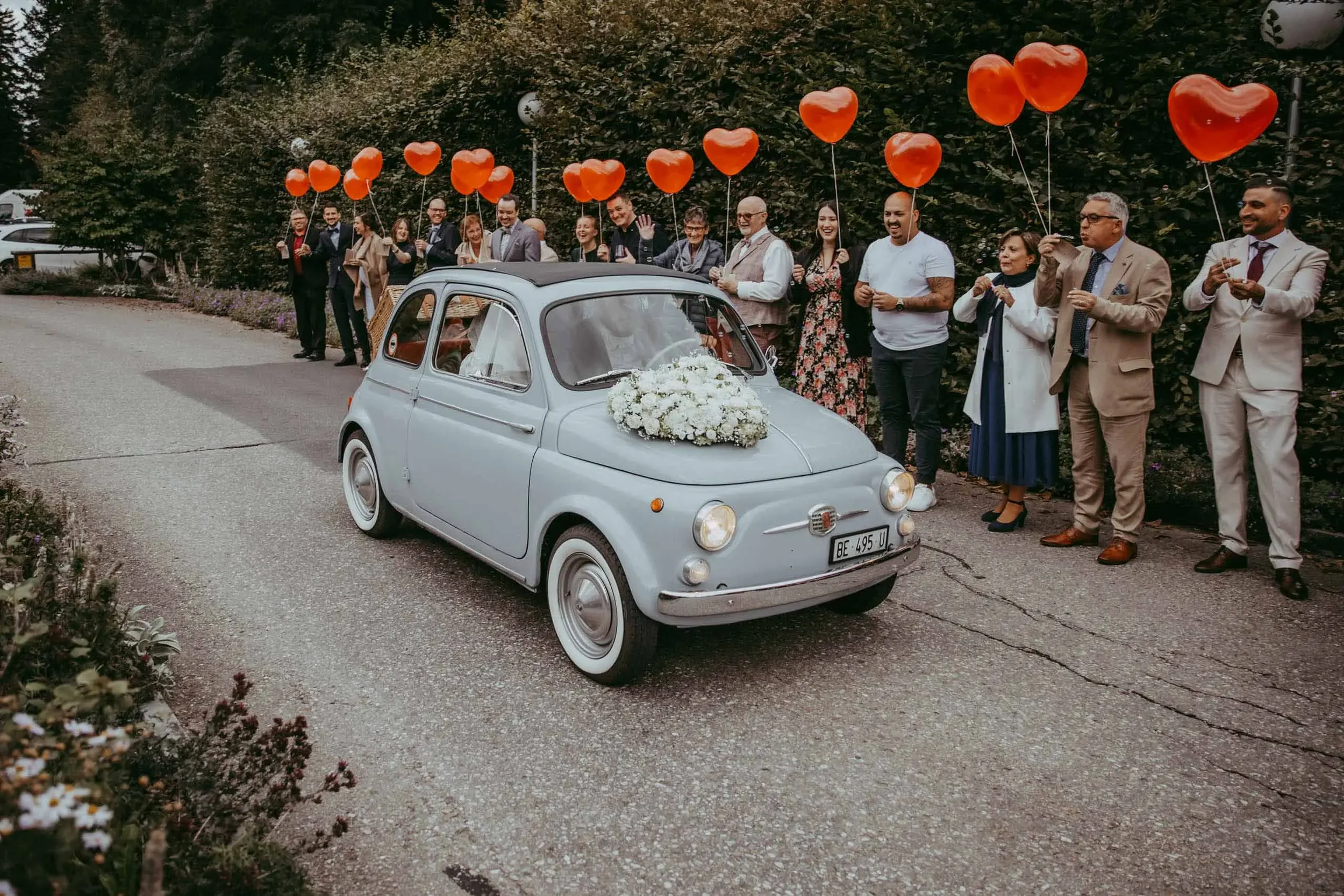 Eine Gruppe von Menschen steht hinter einem kleinen, weißen, klassisch retrofahrzeugartigen Auto mit weißer Blume an der Motorhaube, das mit roten Herzballons verbunden ist. Es sieht nach einer Hochzeit oder Feier aus, im Hintergrund Bäume und ein Pa