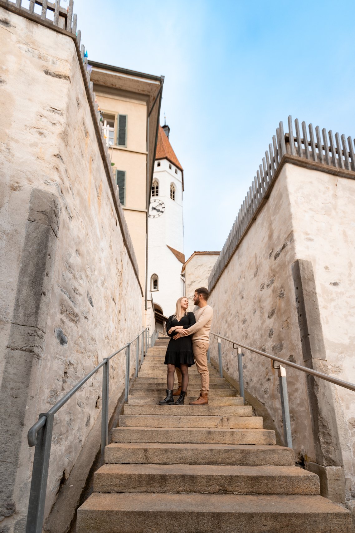 Ein Paar steht auf einer Steintreppe in einer mittelalterlichen Stadt, umgeben von hohen Steinmauern und einer Kirchturm im Hintergrund. Das Paar schaut sich an und lächelt.