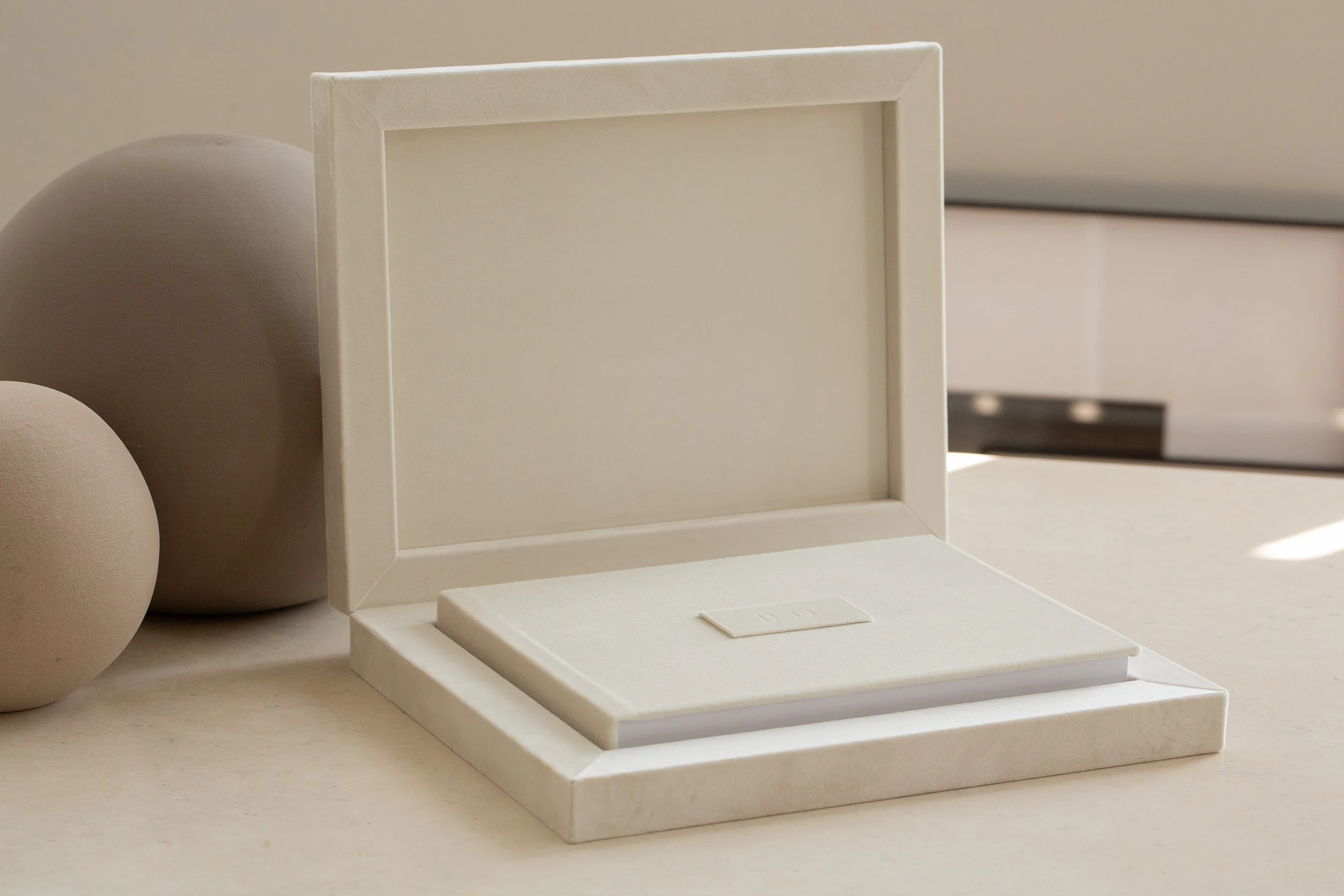 Weiße Schmuckschatulle mit offenem Deckel auf weißem Untergrund, daneben zwei beige Kugeln, im Hintergrund ein grauer Gegenstand.