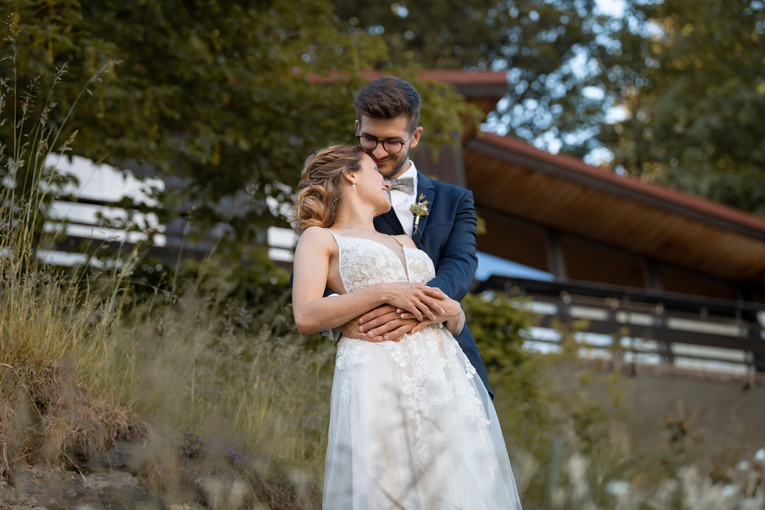 Ein glückliches Paar bei einer Hochzeit im Freien, umarmend und lächelnd. Der Mann trägt einen dunklen Anzug mit Brille, die Frau ein weißes Brautkleid.