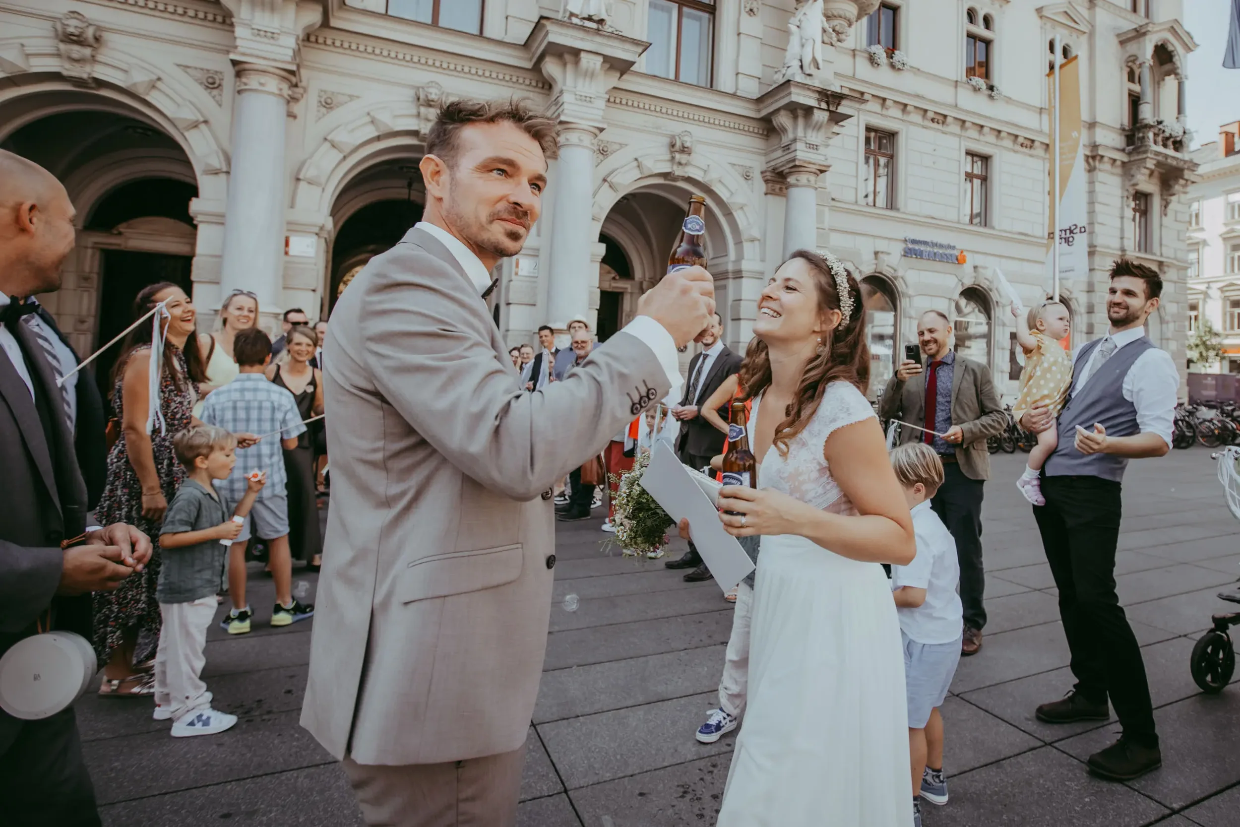 Ein Brautpaar bei ihrer Hochzeit, der Bräutigam schenkt der Braut ein Bier während des Empfangs vor einem historischen Gebäude in der Stadt.