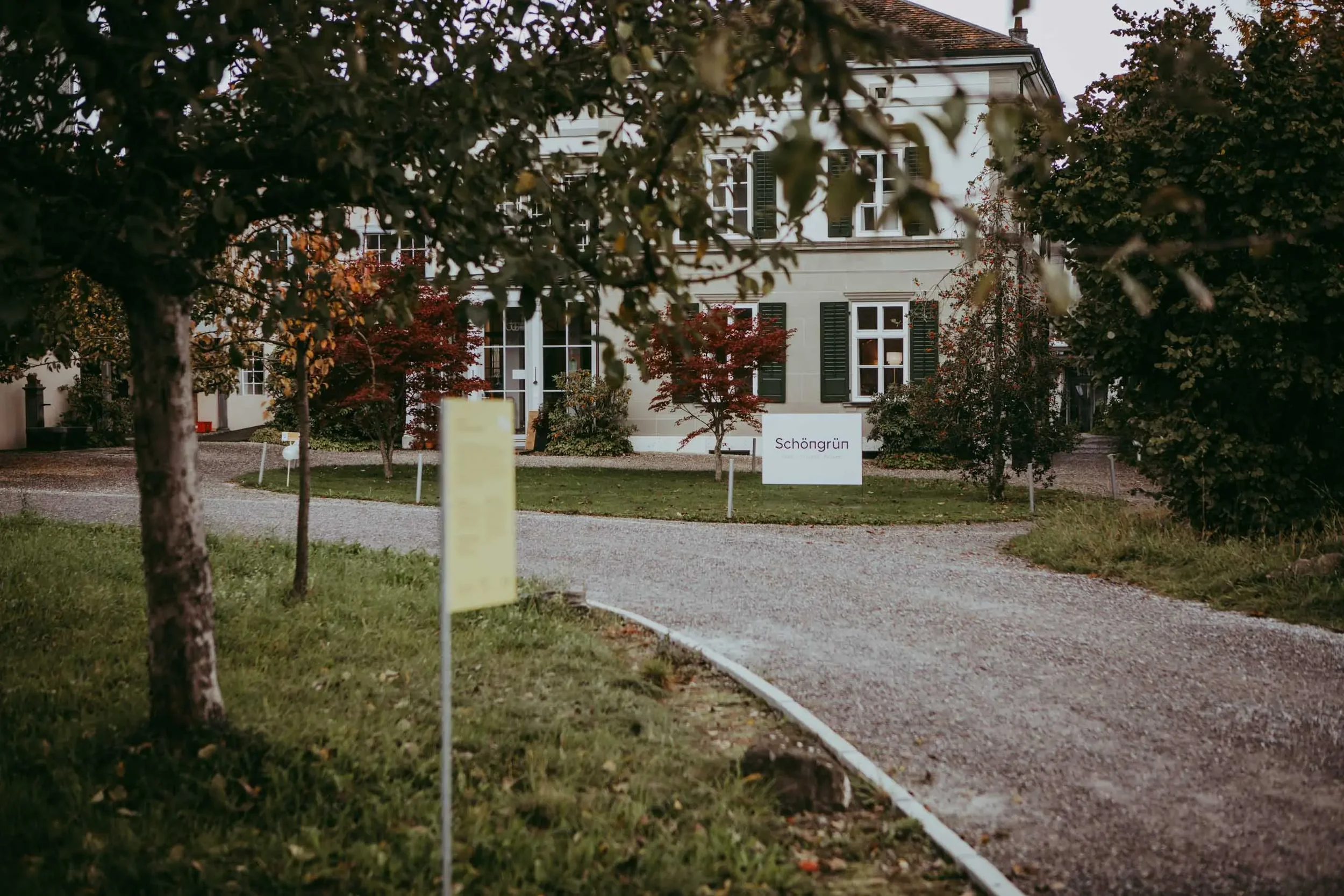 Ein Haus mit mehreren Fenstern und grünen Fensterläden, umgeben von rot- und grünlaubigen Bäumen im Garten. Im Vordergrund eine Vielzahl von Bäumen und eine Allee. Es ist vermutlich Herbst.