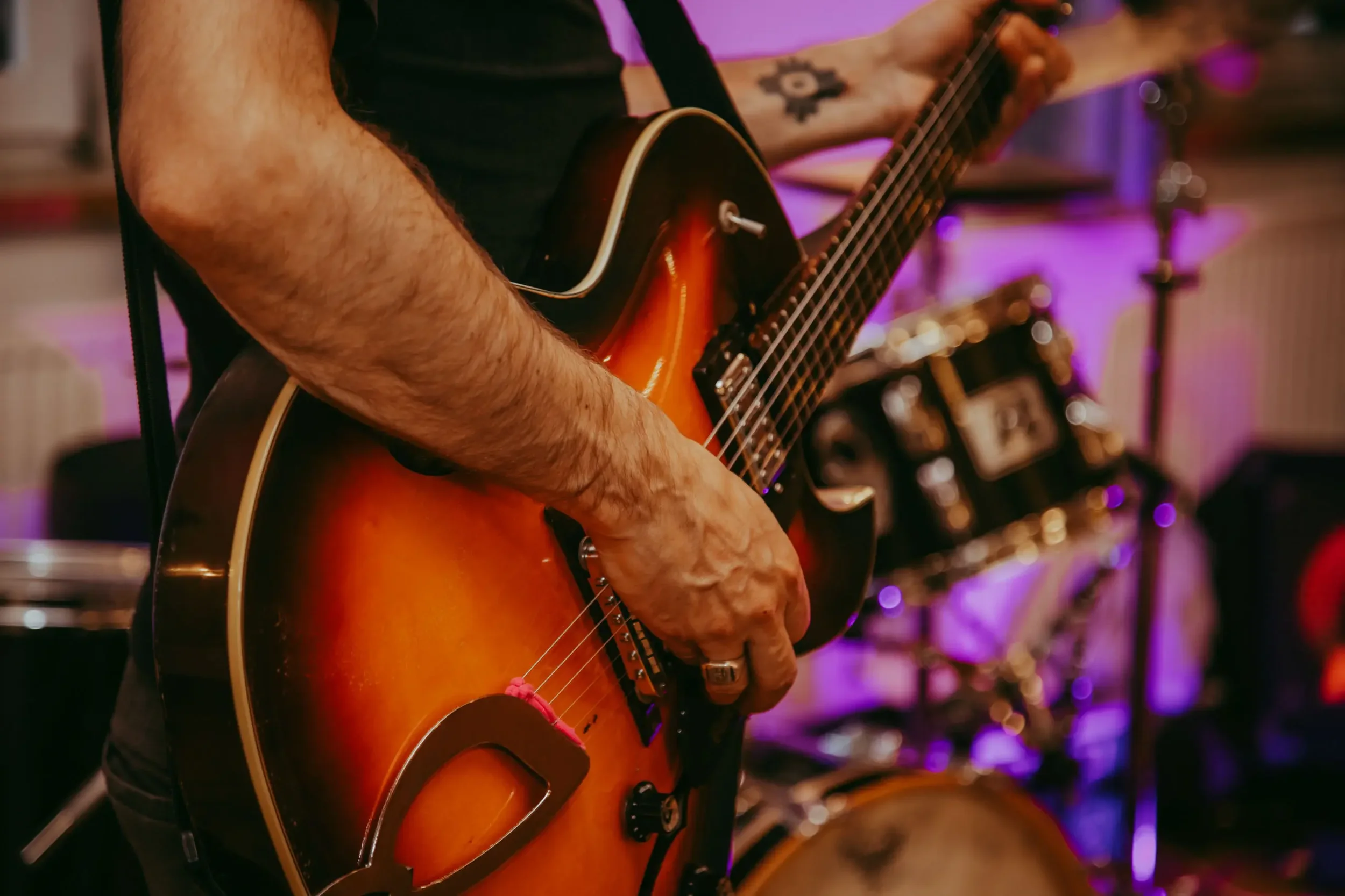 Nahaufnahme eines Mannes, der Gitarre spielt, mit tattoos am Arm, im Hintergrund Instrumente in einem Raum mit buntem Licht.