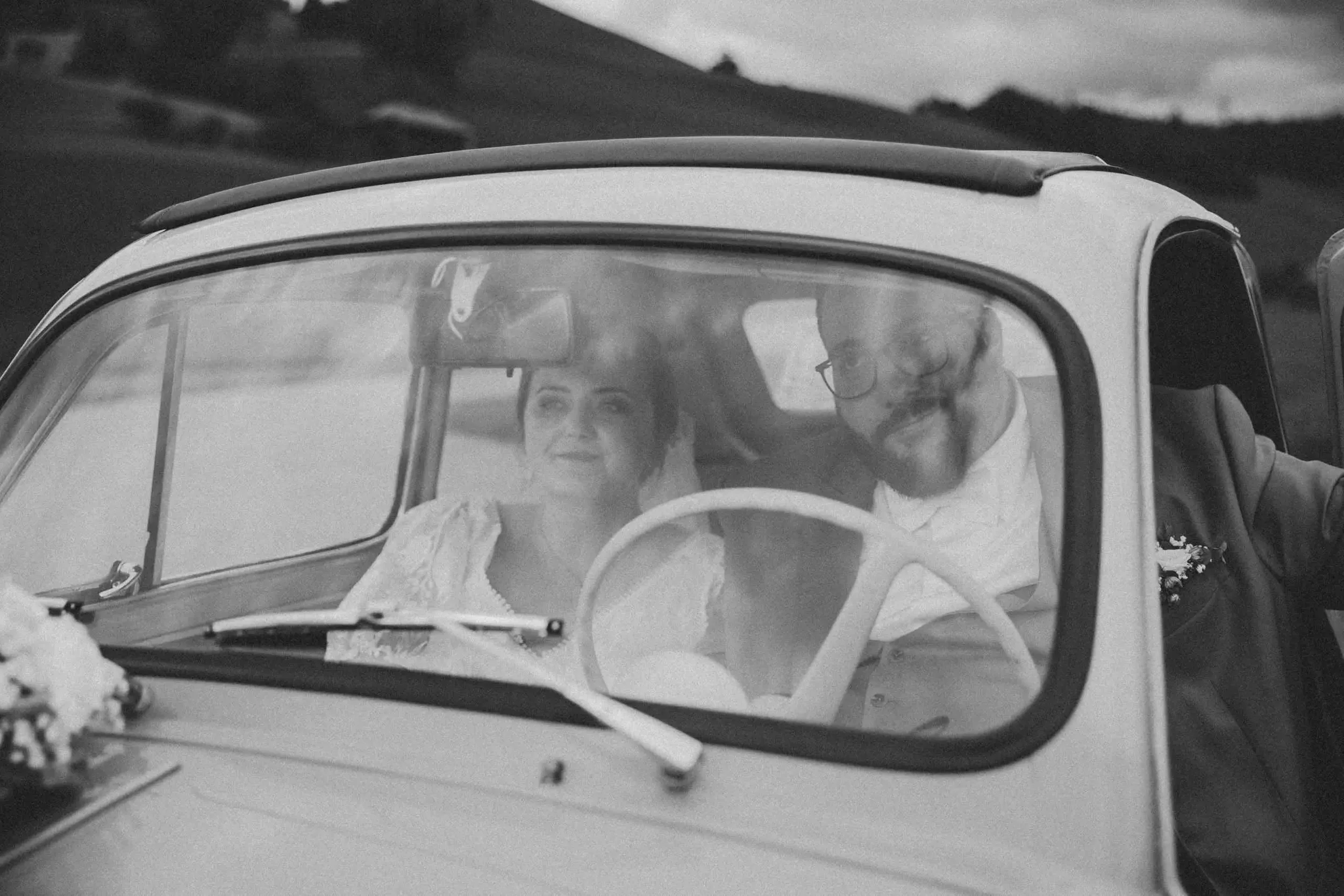Ein schwarzw white Foto eines Paares, das in einem kleinen Auto sitzt, vermutlich an einem besonderen Anlass, mit Omas Blick auf die Straße schauend.