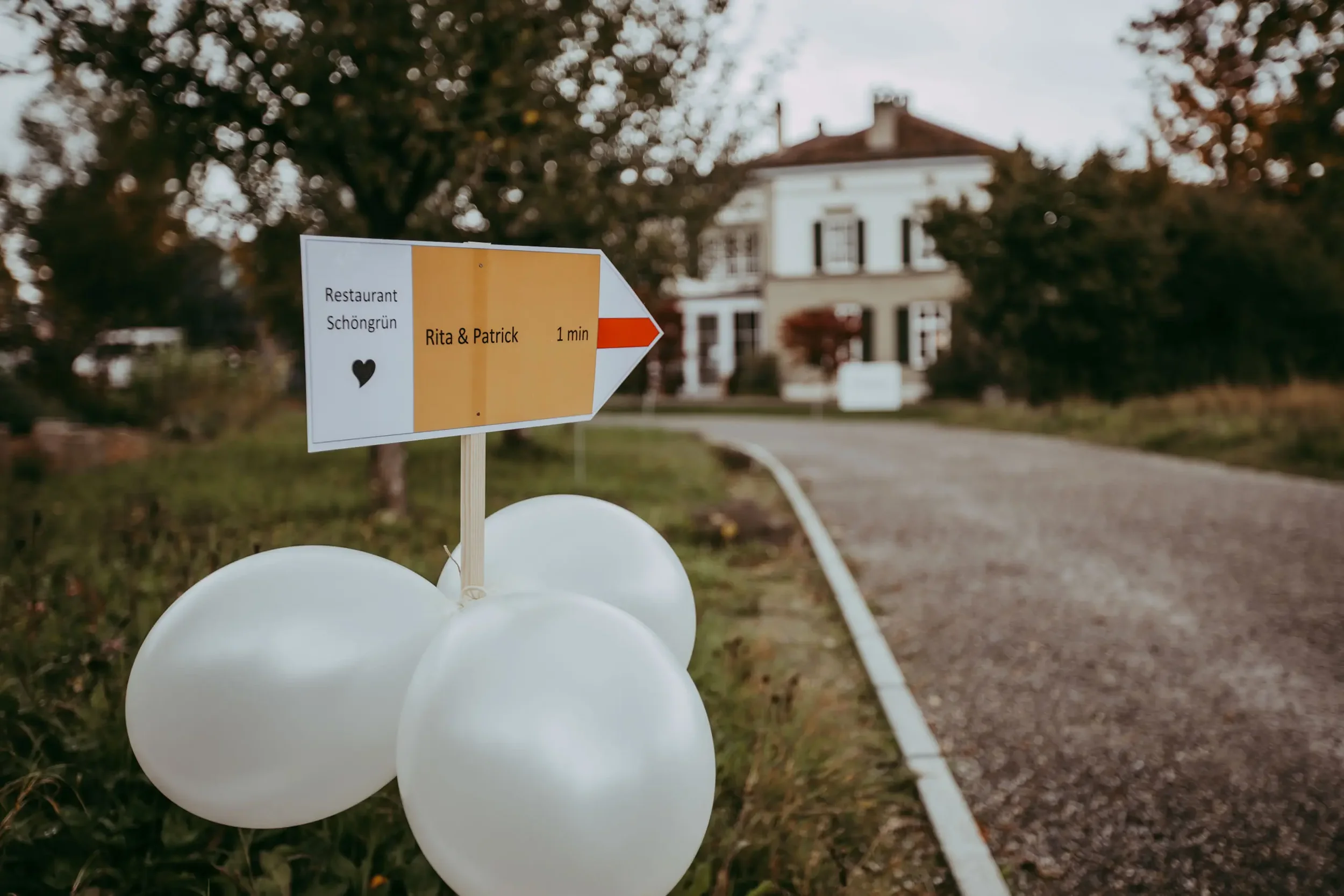 Ein Wegweiser mit der Aufschrift 'Rita & Patrick', der auf ein Restaurant namens Schöngrün hinweist, steht vor einem großen Haus im Garten, mit weißen Ballons am Boden befestigt.