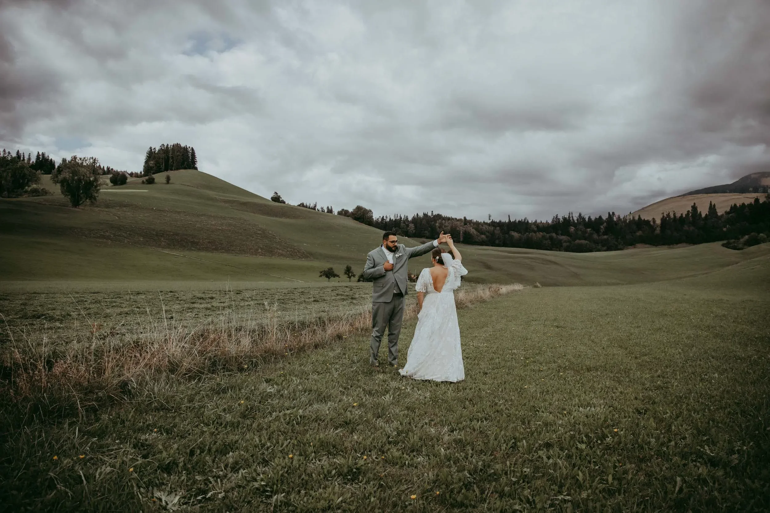 Ein Brautpaar bei ihrer Hochzeit im freien auf einer weiten grünen Wiese, umgeben von Hügeln und Bäumen bei bewölktem Himmel.