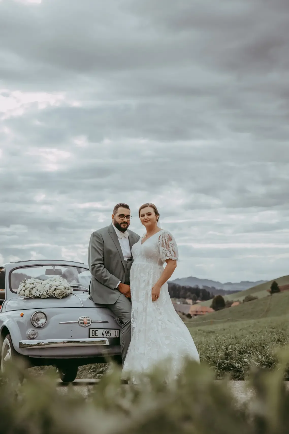 Ein heiratendes Paar steht vor einem grauen Fiat 500 mit Blumen auf der Motorhaube, im ländlichen Gebiet bei bewölktem Himmel.