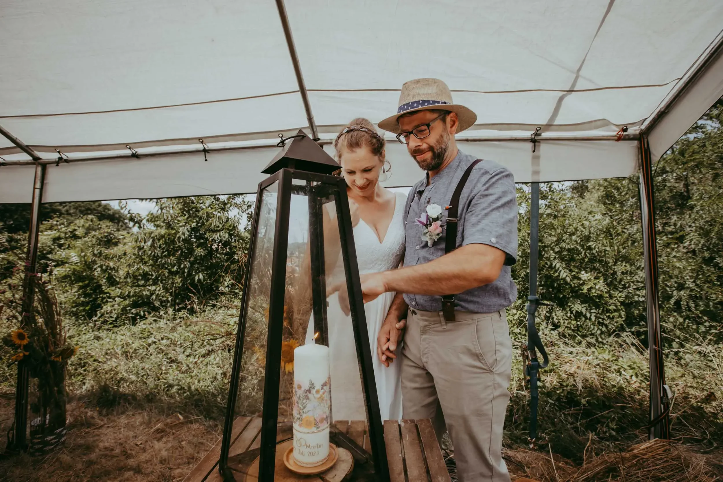 Ein Paar bei einer Hochzeit unter einem Zeltdach. Sie stehen vor einer Taufkerze mit Jubiläumsdaten. Beide lächeln, die Frau trägt ein weißes Kleid, der Mann trägt ein graues Hemd und Hut.