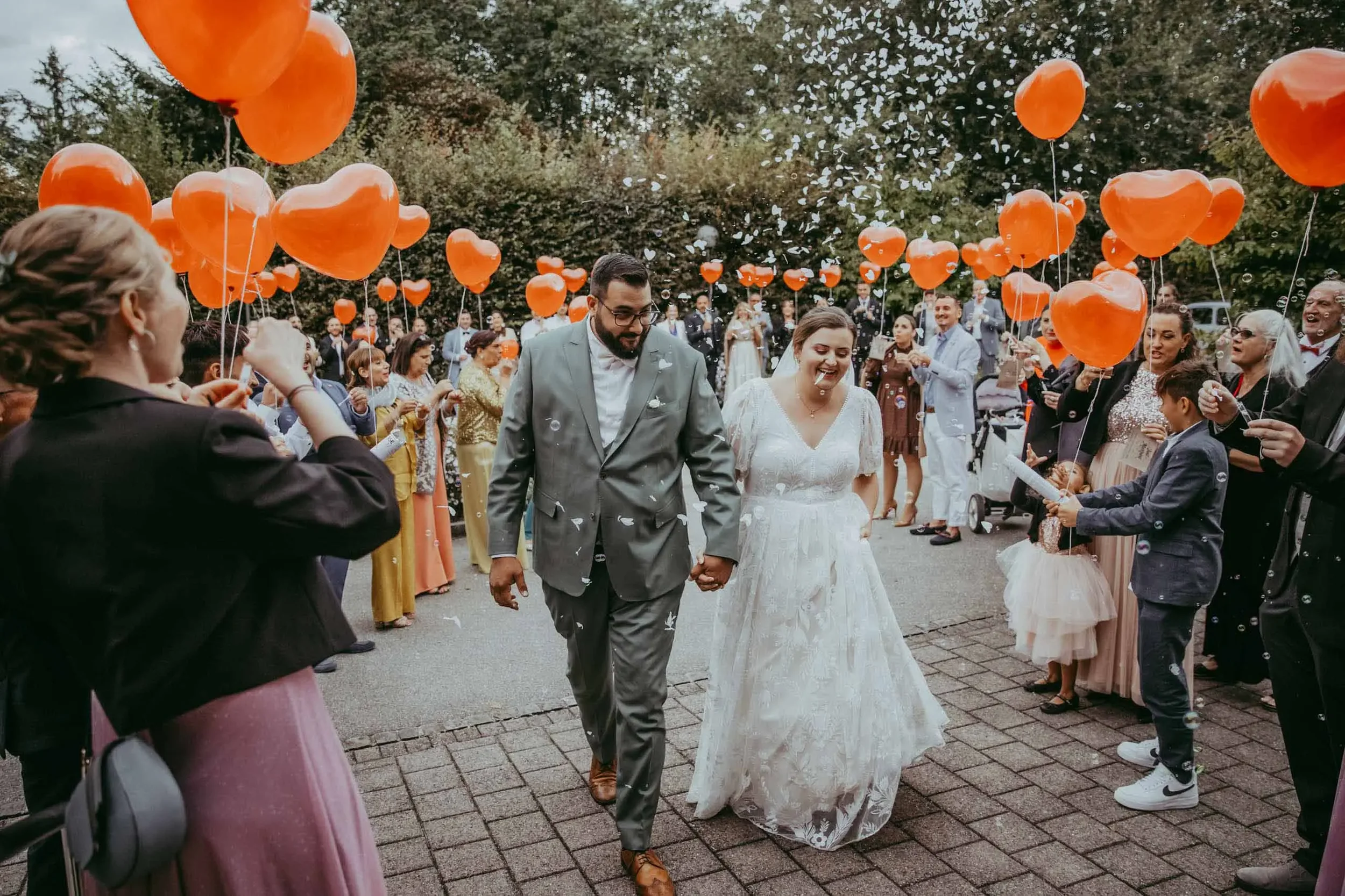 Ein heiratendes Paar, das Hand in Hand geht, umgeben von Freunden und Familie, die Herzballons halten und Seifenblasen blasen, während sie bei einer Gartenhochzeit feiern.