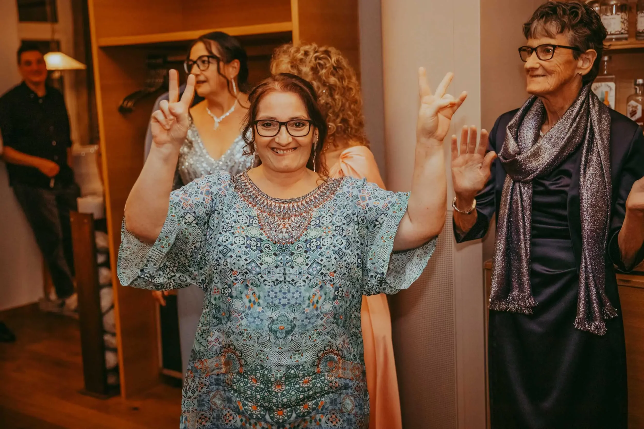 Fünf Menschen, vier Frauen und ein Mann, feiern in einem Wohnzimmer, einige zeigen Peace-Zeichen, alle lachen oder lächeln, im Hintergrund sind Bücherregale und eine Lampe sichtbar.