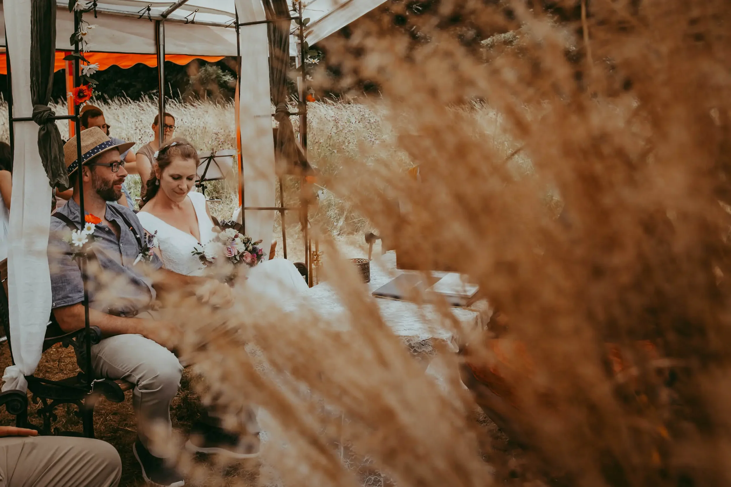 Menschen sitzen bei einer Outdoor-Hochzeit unter einem Gartenpavillon, umgeben von Natur mit trockenen Pflanzen im Vordergrund.