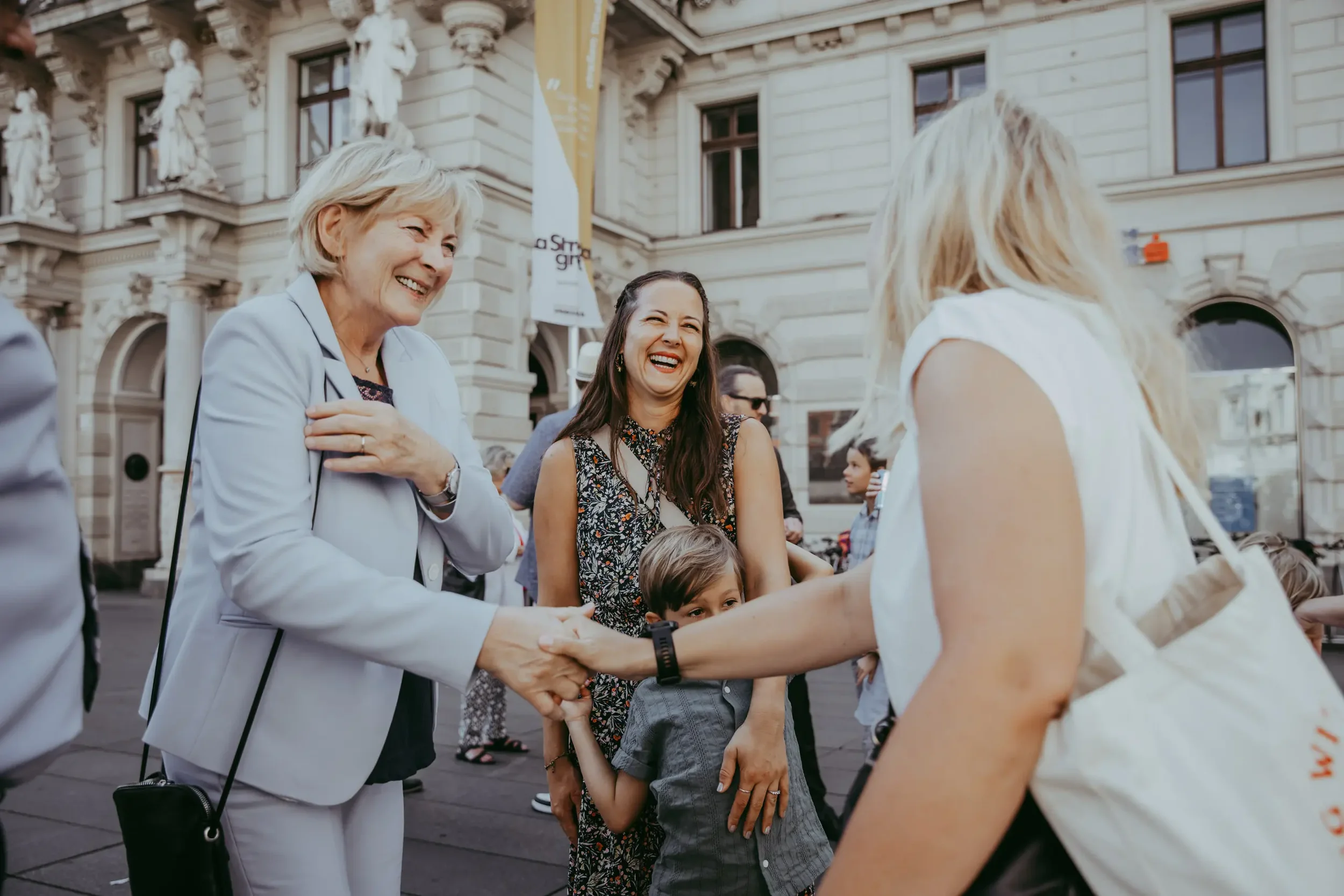 Drei Frauen und zwei Kinder beim Händeschütteln vor einem historischen Gebäude, lächelnd bei einer Veranstaltung in der Stadt, im Freien.