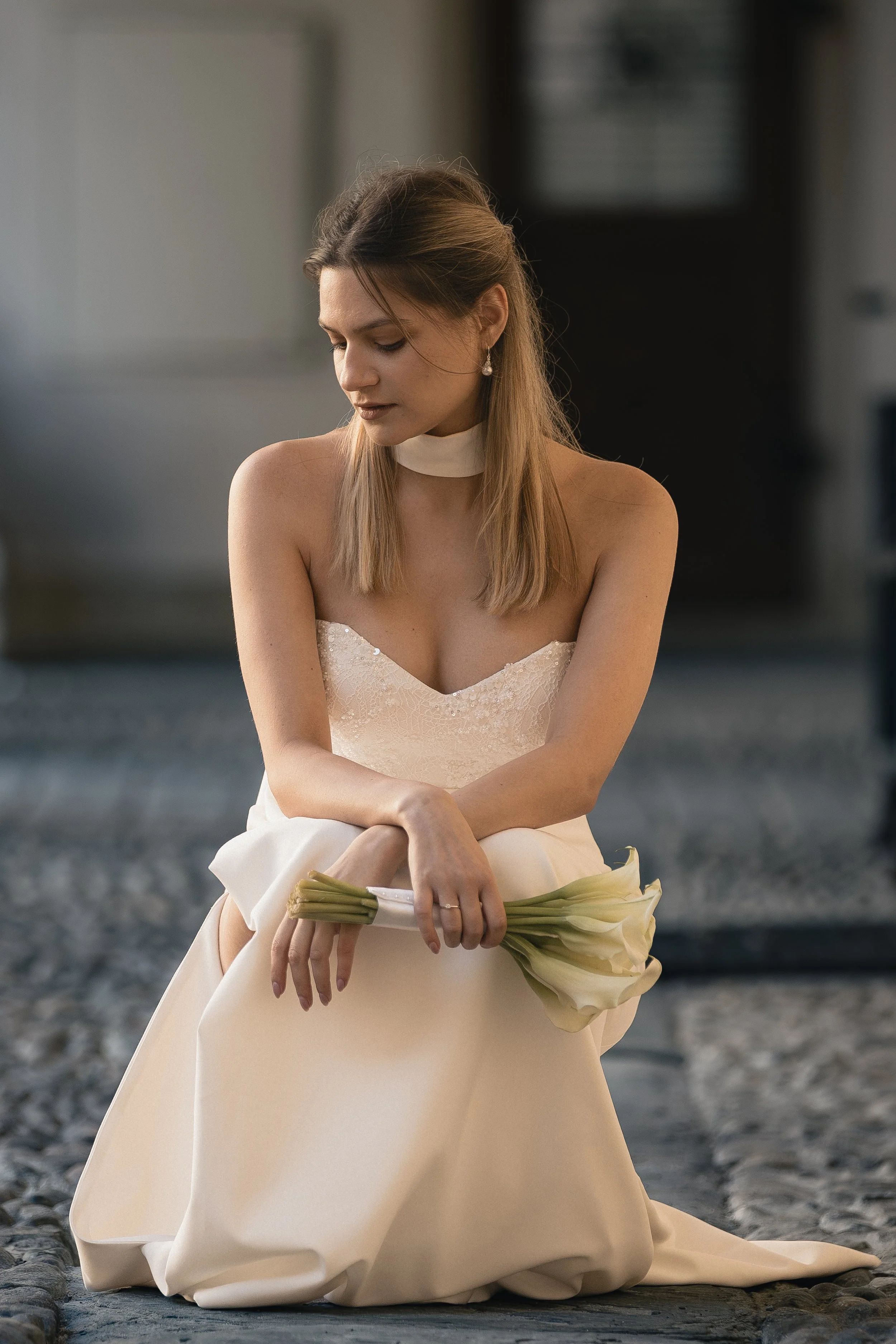Frau in weißem Brautkleid sitzt auf einem Steinweg, hält einen Blumenstrauß und blickt nach unten.