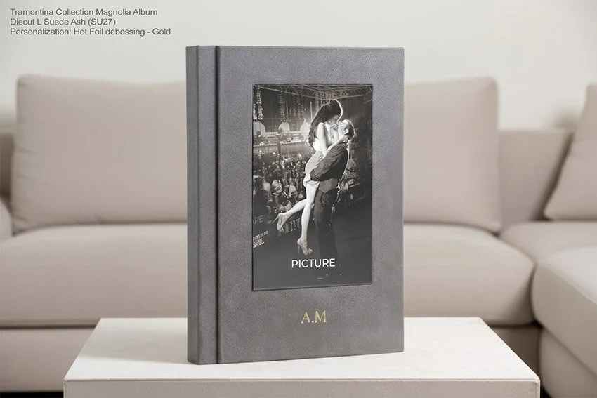 Ein graues Fotobuch auf einem weißen Tisch, das eine Schwarz-Weiß-Fotografien von einem Mann und einer Frau in einer Umarmung zeigt, in einem Wohnzimmer mit grauen Sofas im Hintergrund.