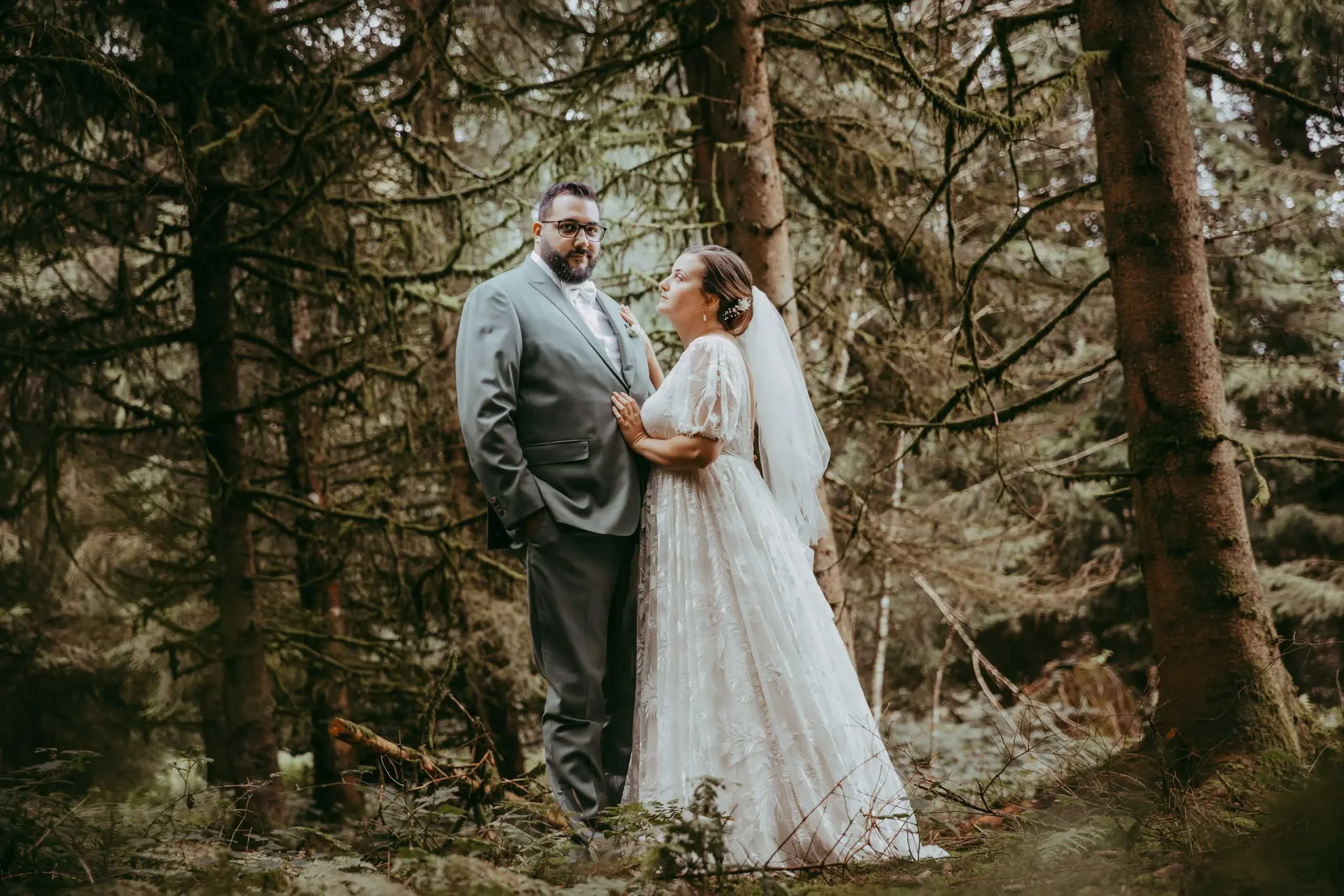 Ein Brautpaar im Wald während einer Hochzeitsfotografie, der Mann trägt einen grauen Anzug, die Frau ein weißes Hochzeitskleid mit Schleier.