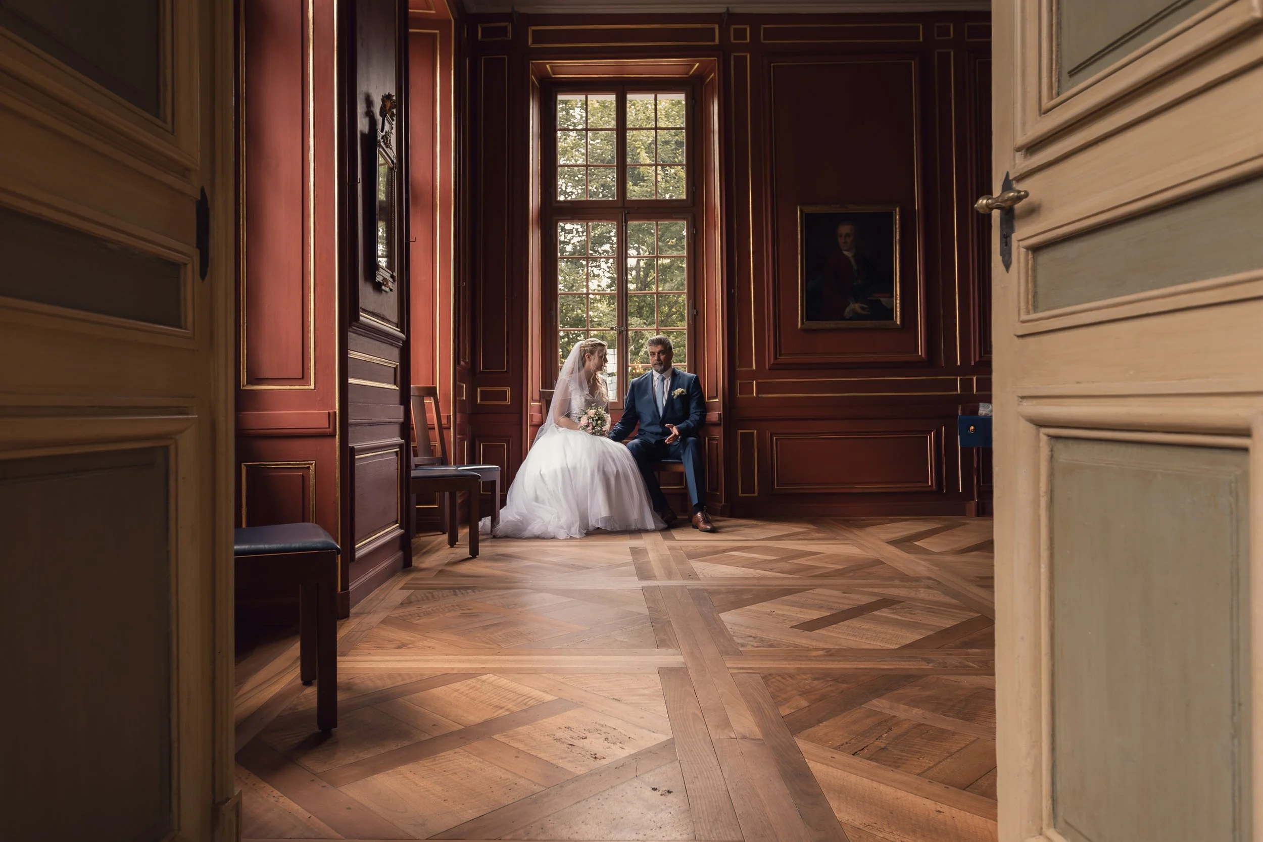 Braut und Bräutigam sitzen in einem eleganten Raum mit großen Fenstern und dunklen Holzwänden, es sieht nach einer Hochzeitszeremonie aus.