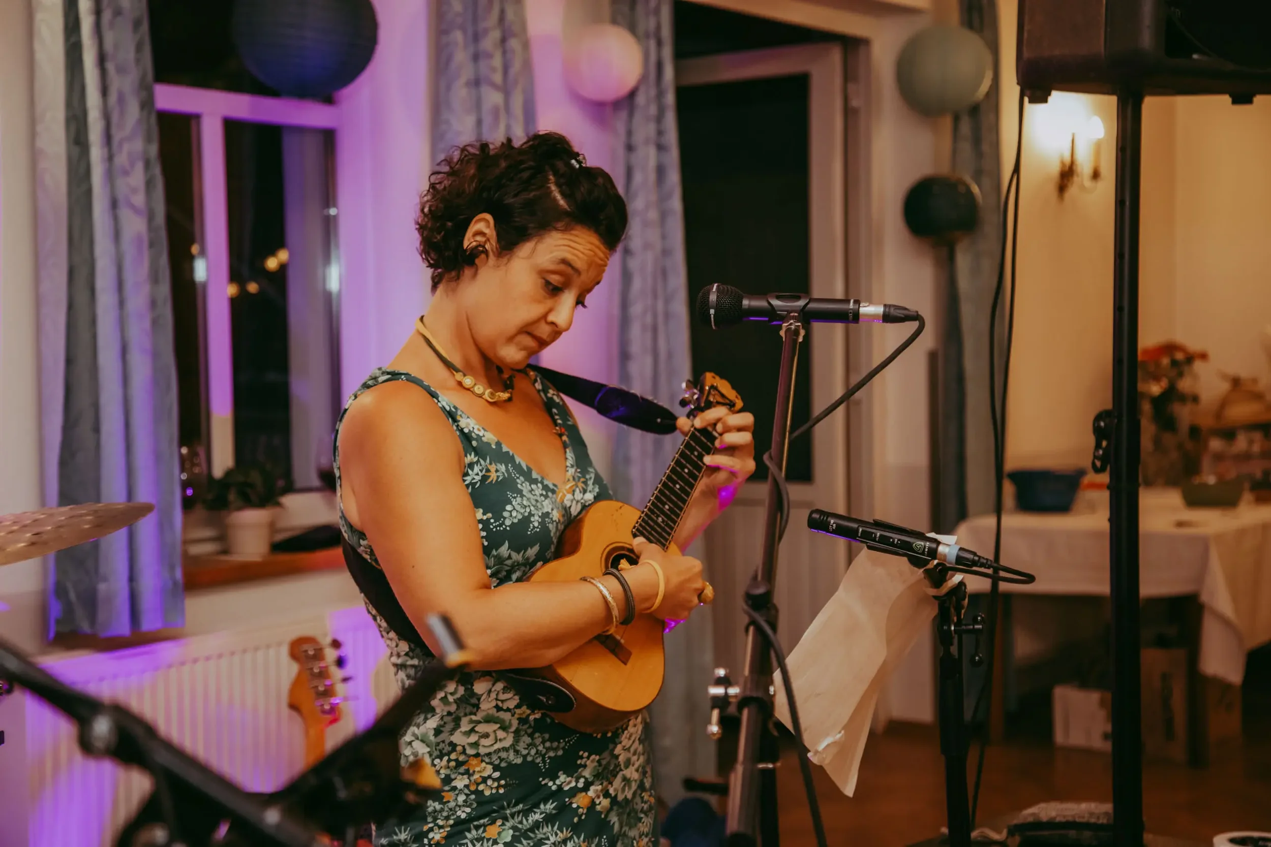 Eine Frau spielt Gitarre in einem gemütlichen Raum mit Laternen und Fenster im Hintergrund, bei einer musikalischen Darbietung.