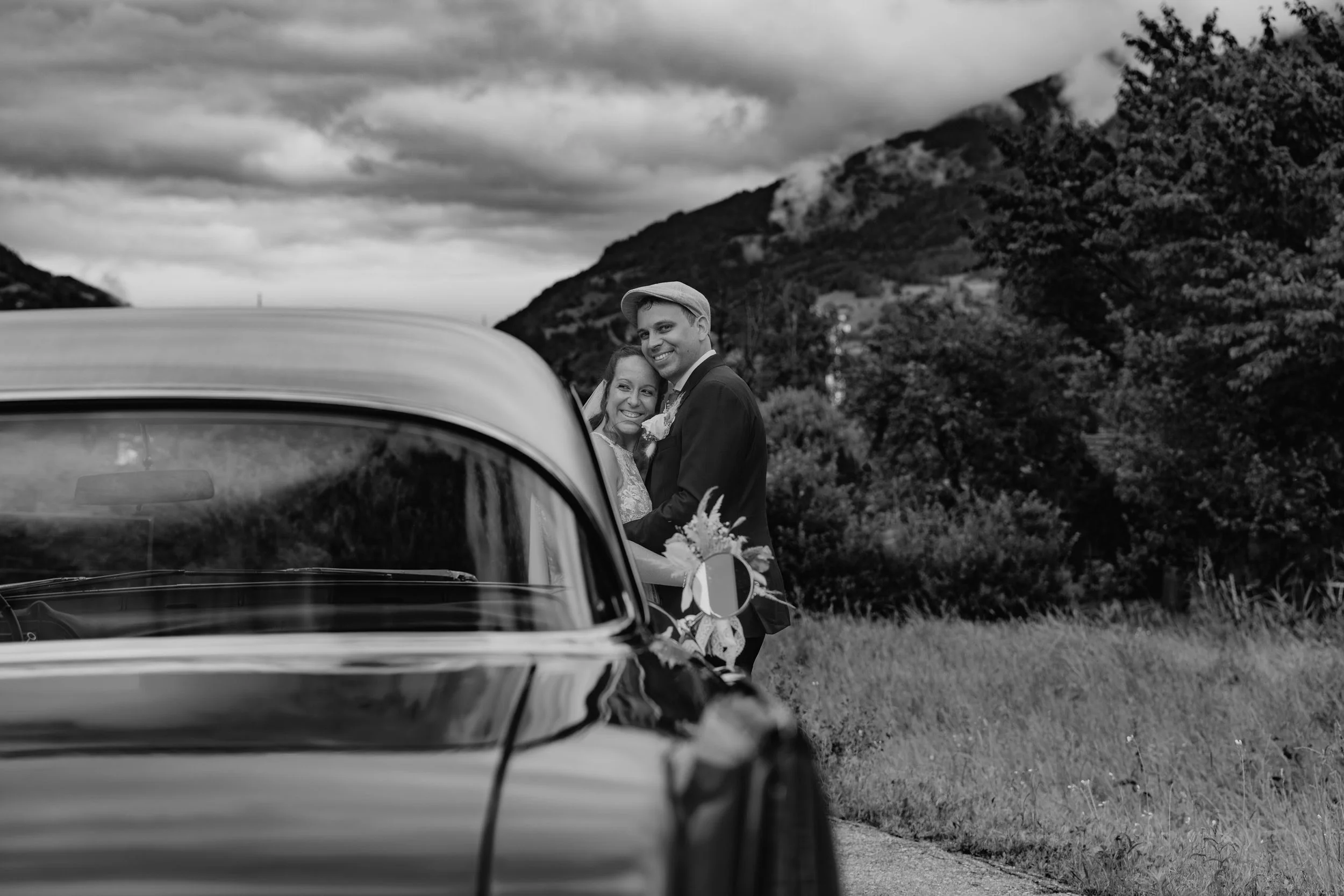 Neues Ehepaar steht vor einem Auto, umgeben von Bergen und Bäumen, bei bewölktem Himmel. Schwarz-Weiß-Fotografie.