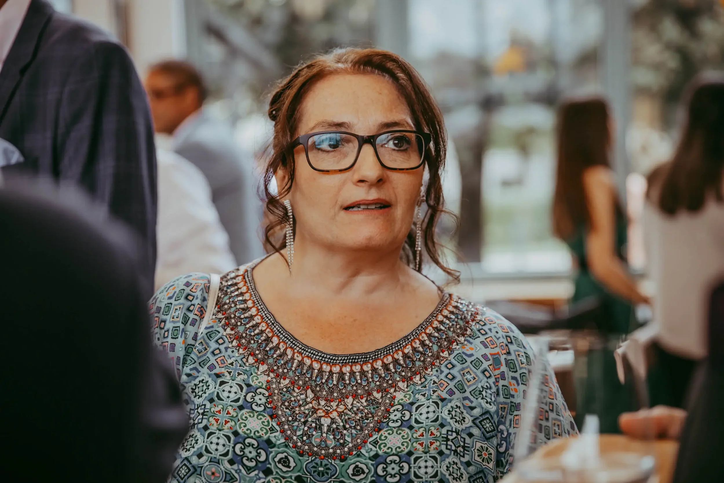 Frau mit dunklen, lockigen Haaren und Brille, trägt ein bunt gemustertes Kleid mit aufwändiger Stickerei, vermutlich bei einem gesellschaftlichen Anlass in einem Raum mit großen Fenstern.