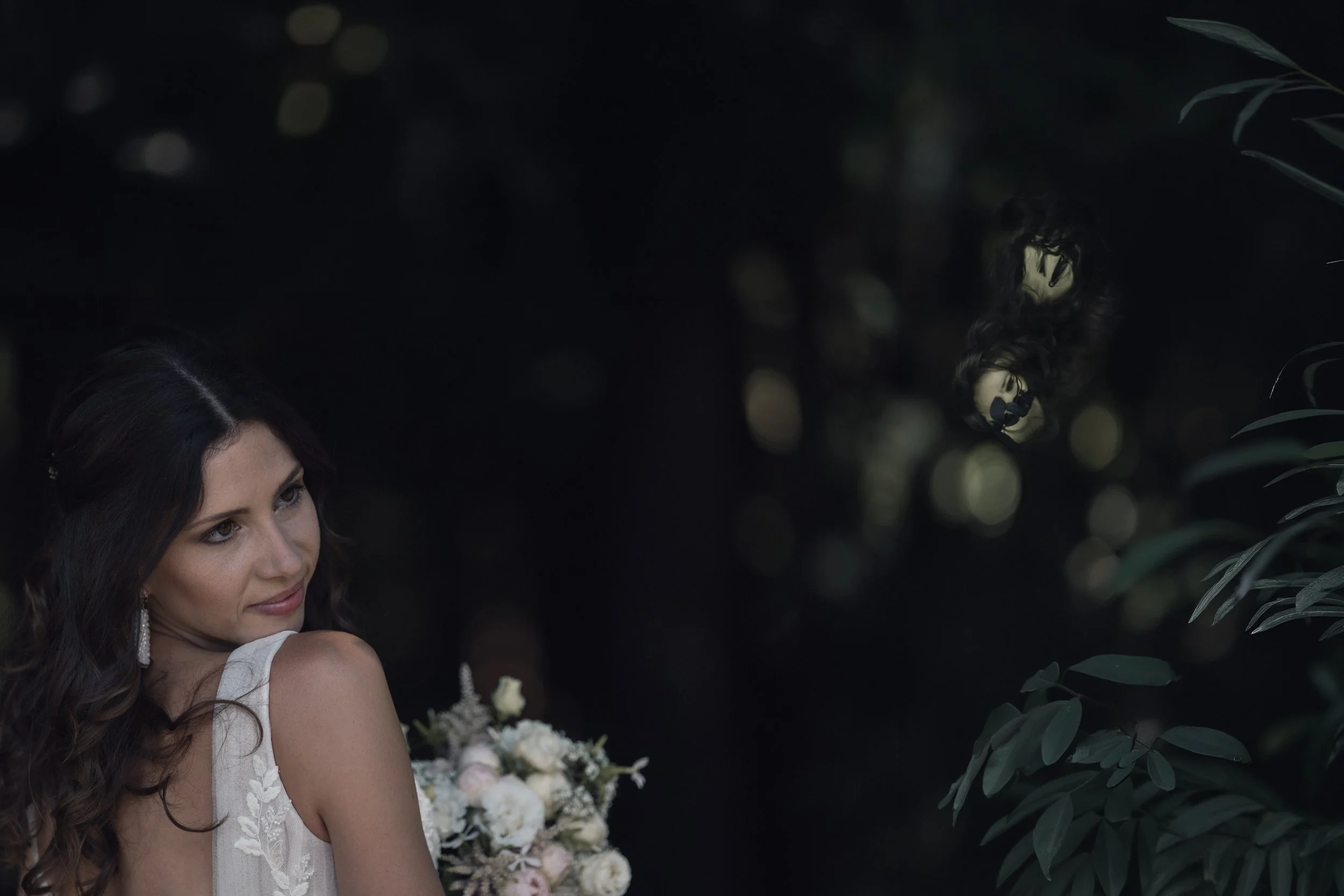 Eine Frau mit dunklen, lockigen Haaren trägt ein weißes Kleid und hält einen Blumenstrauß, während sie im Freien steht, umgeben von grünen Blättern und Bokeh-Lichtern im Hintergrund.