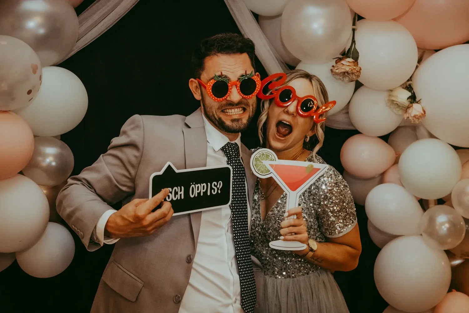 Zwei Hochzeitsgäste mit lustigen Brillen und Requisiten vor einem Ballonhintergrund