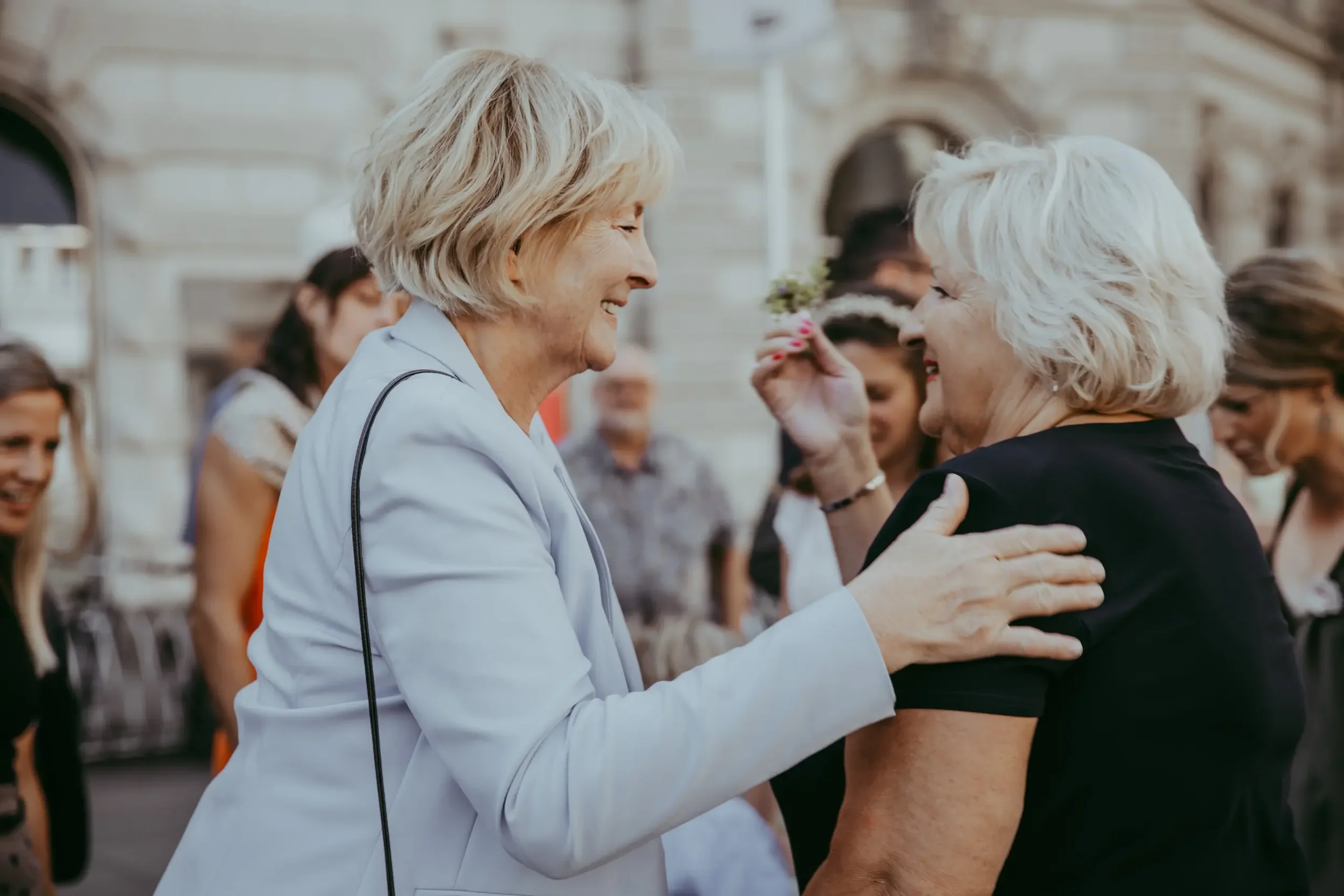 Zwei ältere Frauen beim Händeschütteln und umarmen sich in einer Menschenmenge im Freien, im Hintergrund sind andere Personen. Es scheint eine freundliche Begrüßung oder Zusammenkunft zu sein.