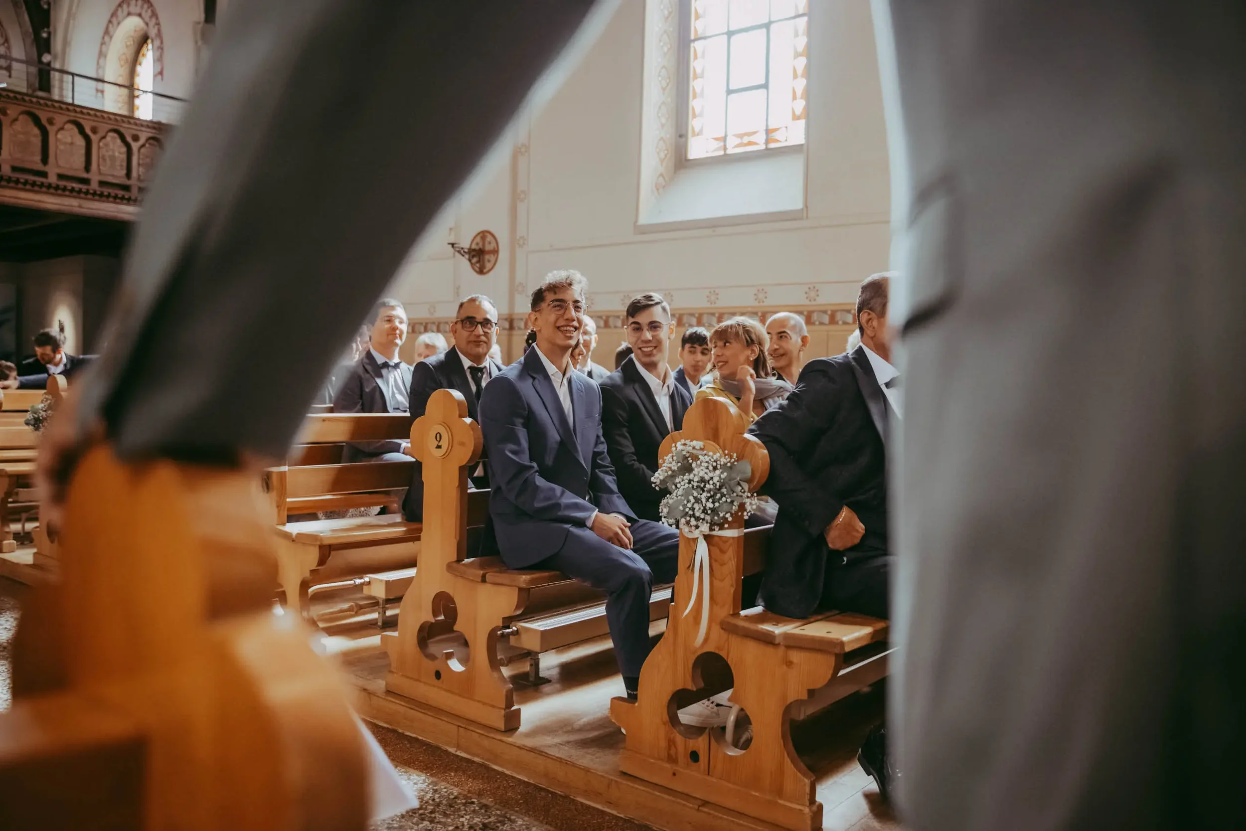 Menschen sitzen in einer Kirche während einer Zeremonie, lachend und lächelnd, sichtbare Holzbank und bunte Fenster im Hintergrund.