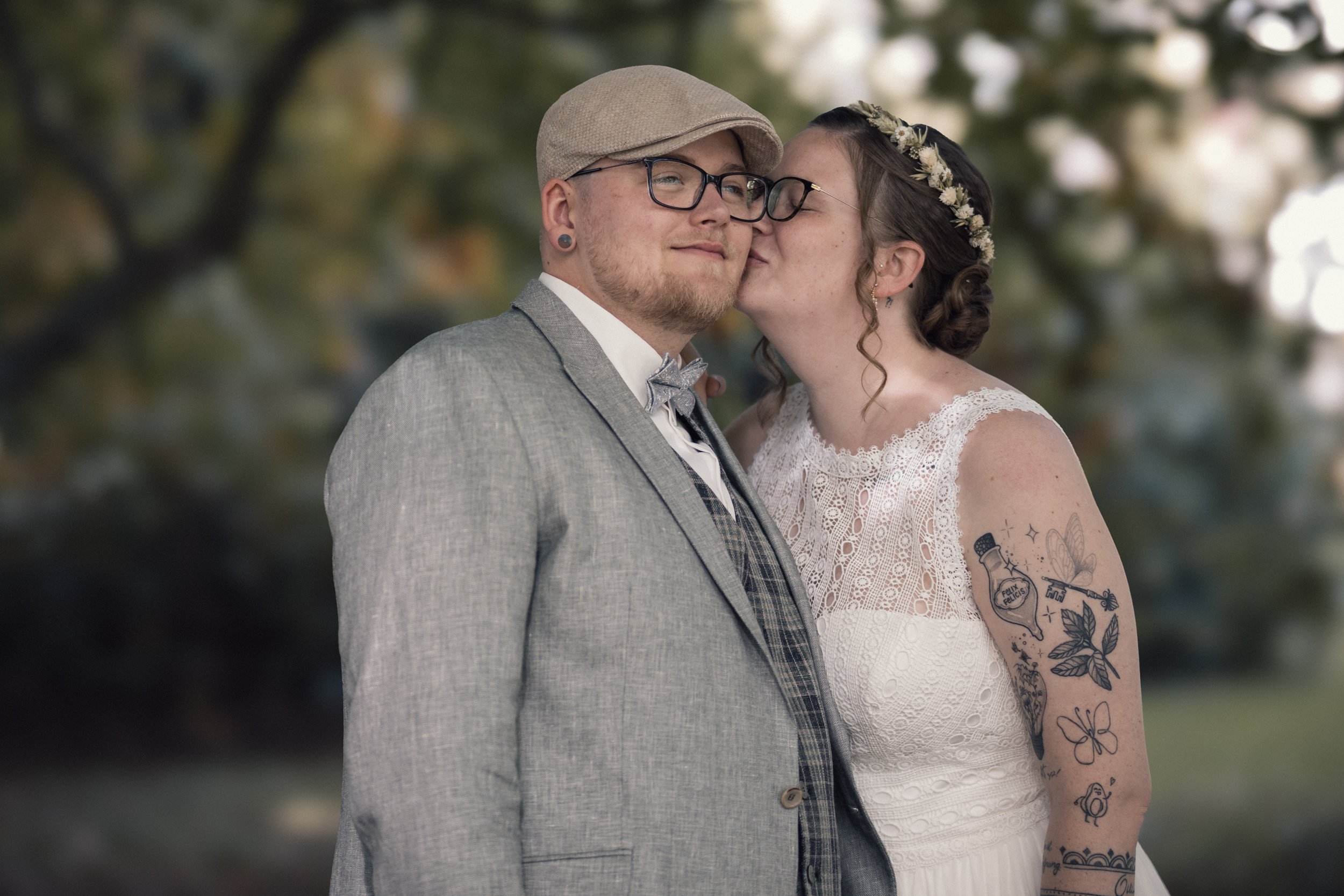 Paar bei einer Hochzeit, der Mann trägt eine Brille, einen grauen Anzug und eine Kappe, die Frau trägt eine weiße Hochzeitstracht mit Tattoos am Arm und eine Blumenkrone im Haar, sie küssen sich.