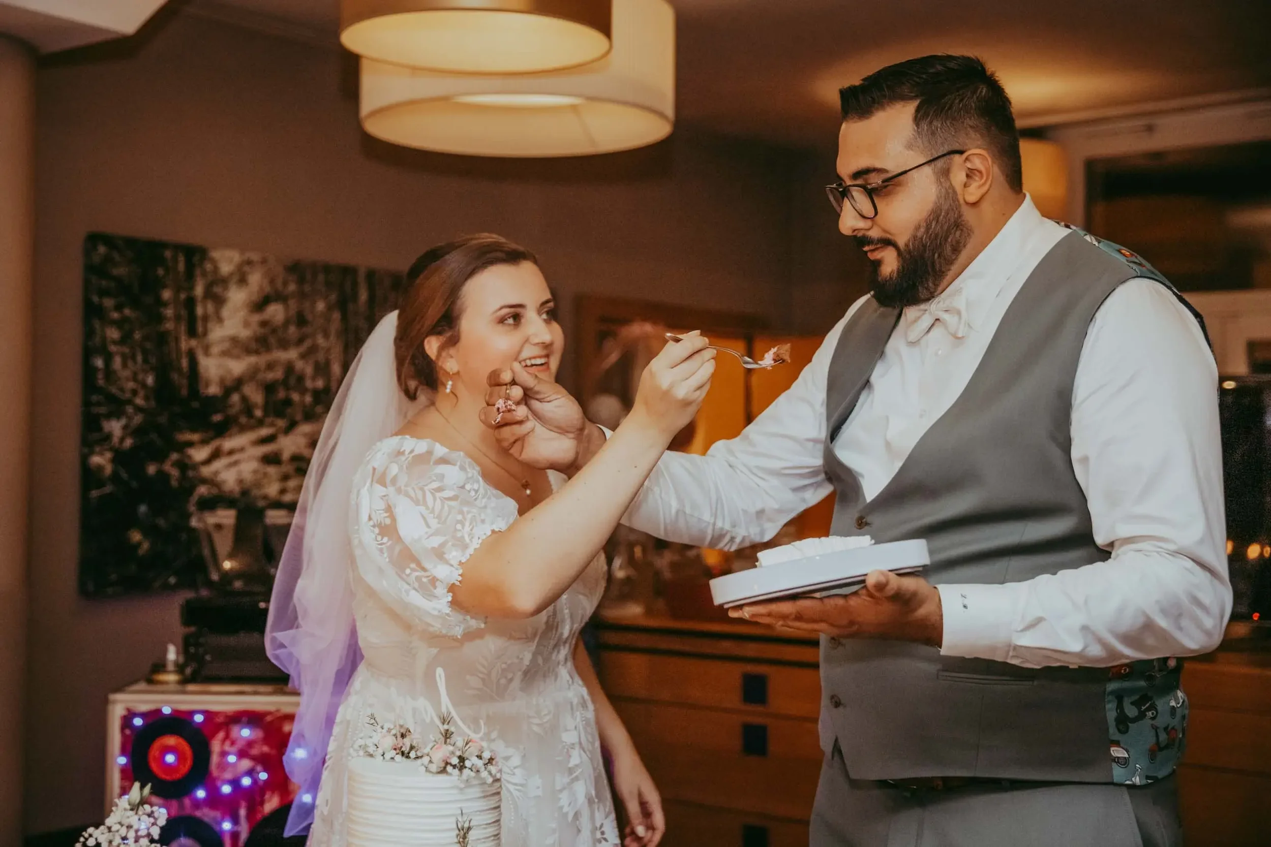 Ein Brautpaar bei einer Hochzeitsfeier, die Braut trägt ein weißes Kleid mit Spitzen und einen Schleier, der Bräutigam trägt einen grauen Anzug. Sie scheinen sich auf einen besonderen Moment zu freuen, während er ihr etwas zu essen auf einen Löffel g