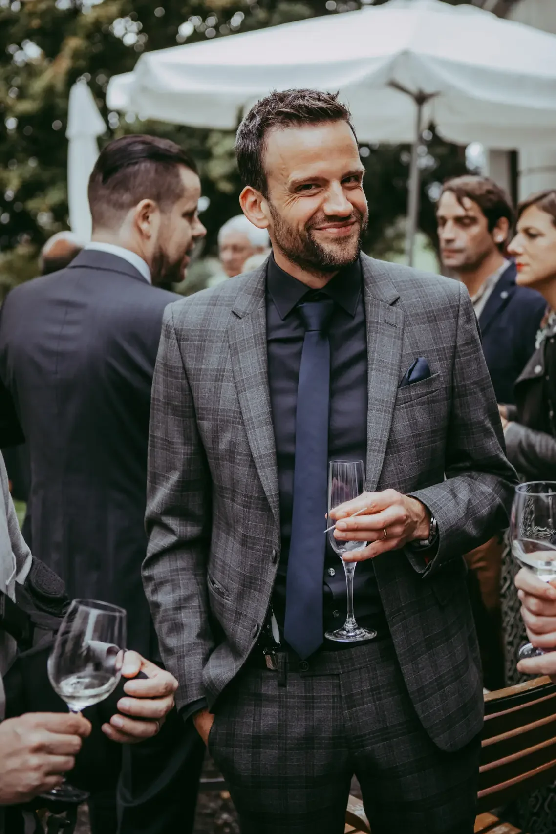 Ein Mann trägt einen grauen Anzug mit roten Linien, hält ein Glas Sekt und lächelt bei einer gesellschaftlichen Veranstaltung im Freien.